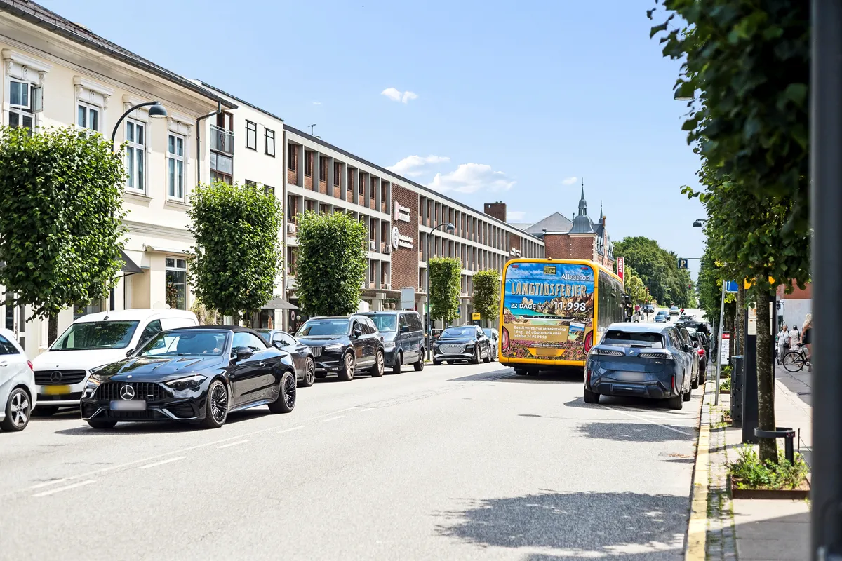 Menighedsgården i Charlottenlund med flere biler parkeret langs gaden og en bus kører forbi