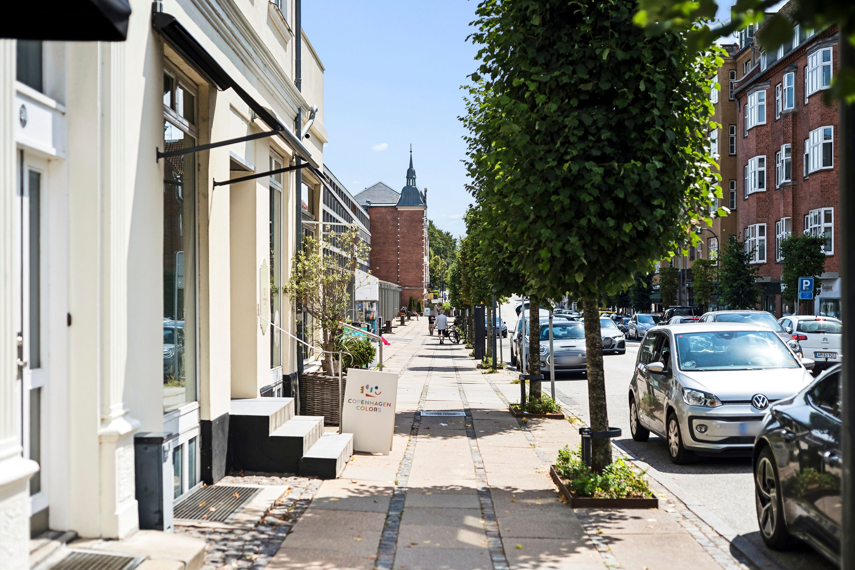 Butik til leje i Charlottenlund. Fortov med parkerede biler og grønne træer. Solrigt vejret og eksklusivt handelsområde.