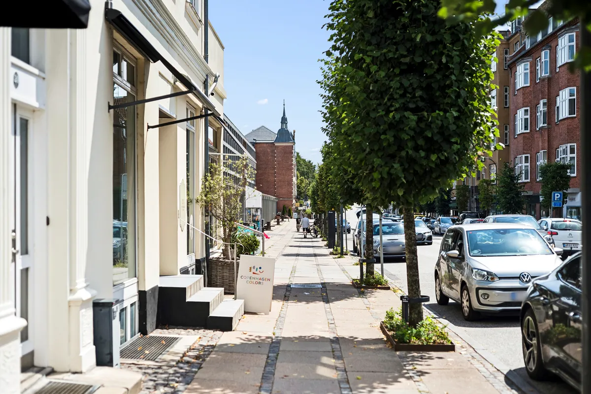 Butik til leje i Charlottenlund. Fortov med parkerede biler og grønne træer. Solrigt vejret og eksklusivt handelsområde.