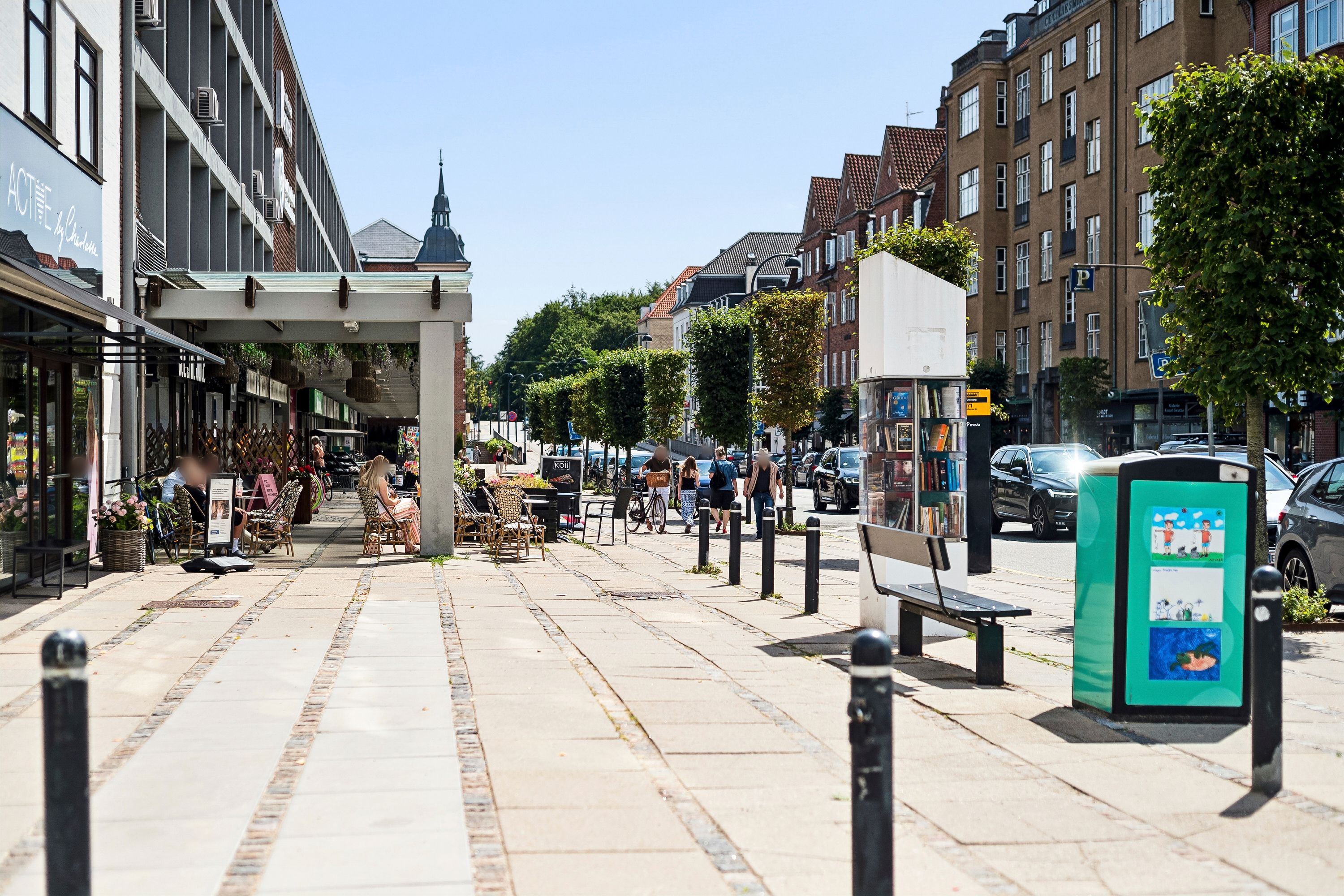 Butiksgade med folk, bænke, parkerede biler, grønne planter og træer, gult skilt, hus og bygning, lyse himmel og vej med fortov