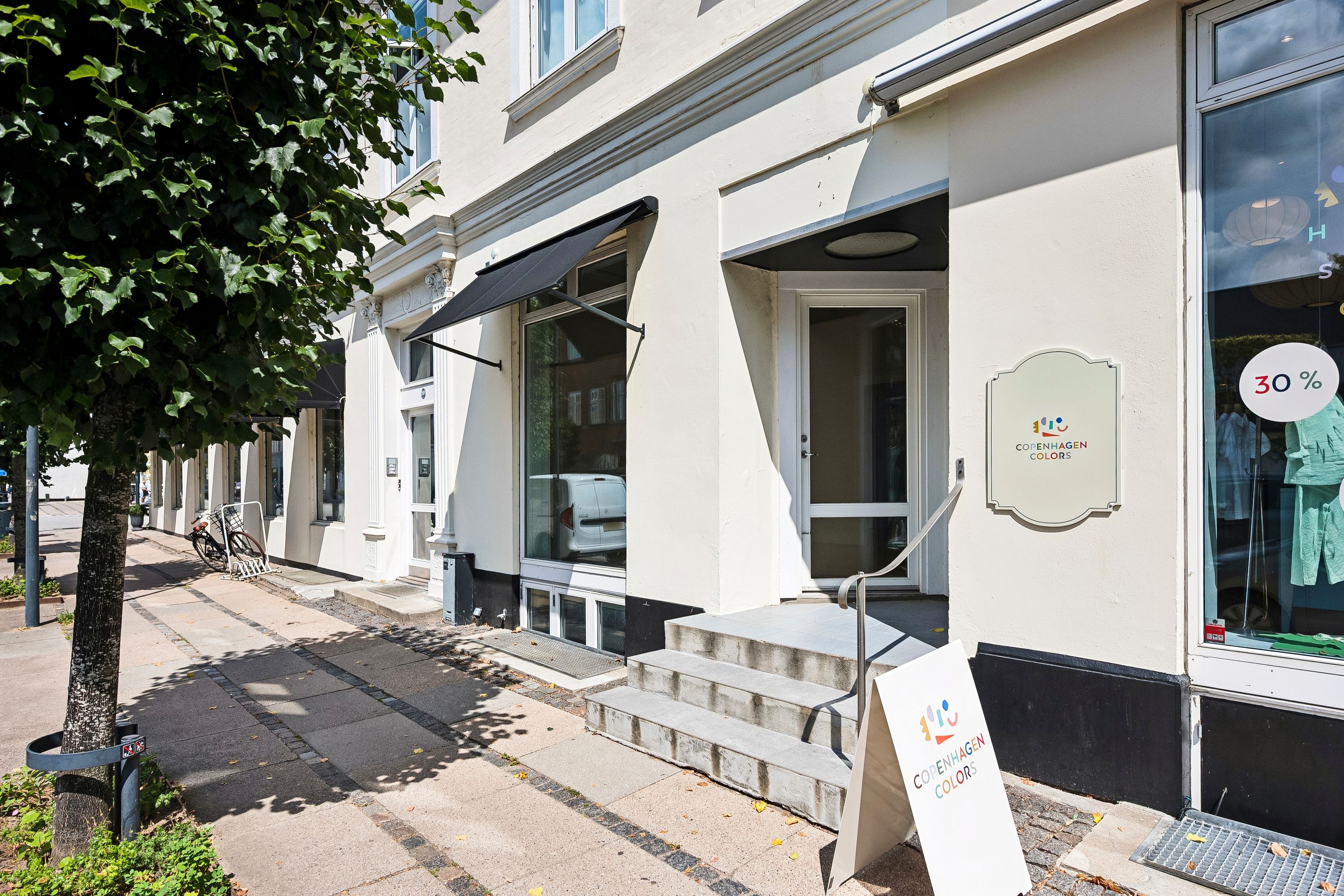 Butiksforretning med mærkat, skilt og vindue med 30 % rabat. Foran vinduet er der en række cykler parkeret, og der er et grønt træ i forgrunden