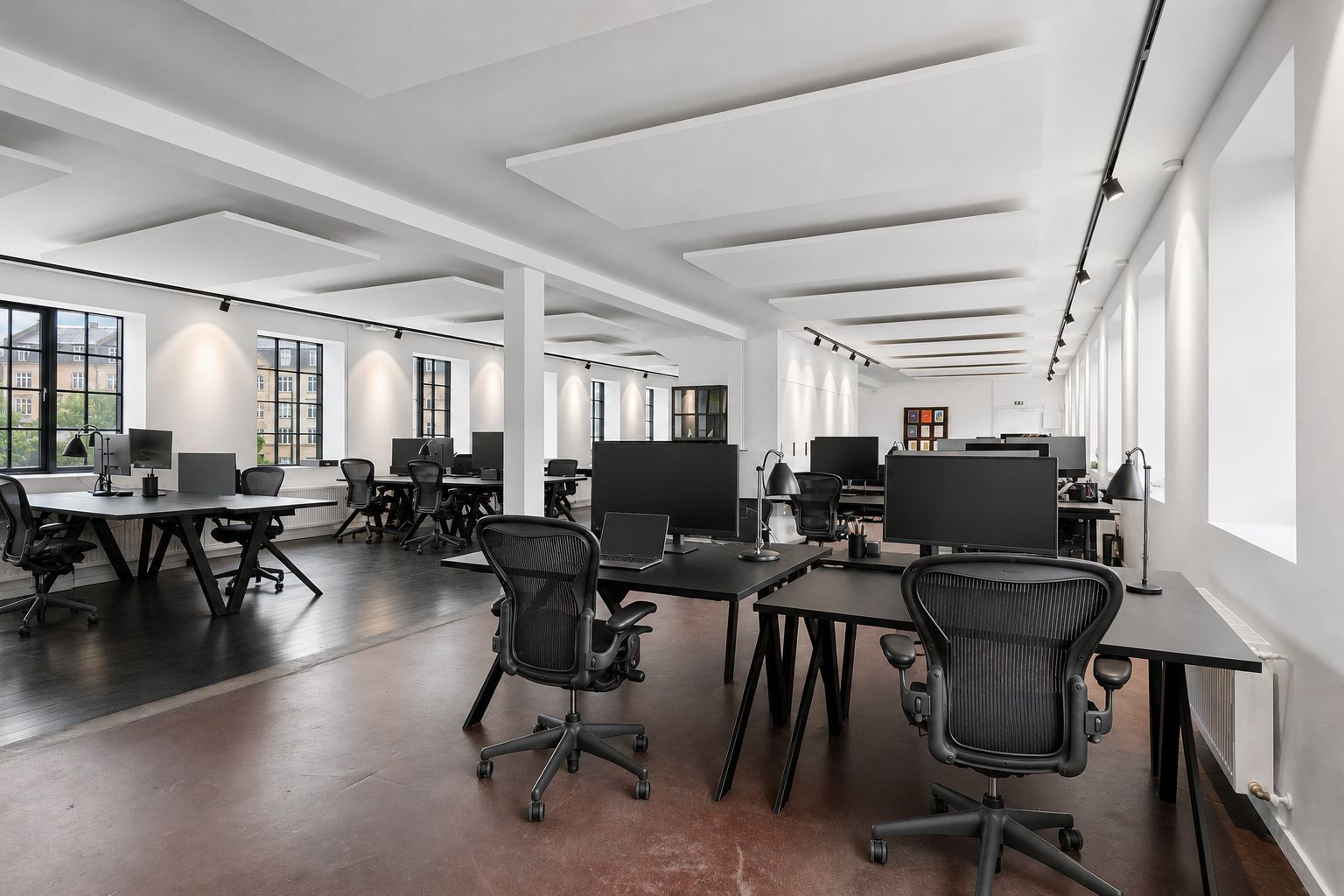 Kontorlokaler med lyse ruder, højt til loftet, og moderne møbler. Sorte borde og skriveborde med monitorer, og ergonomiske stole.