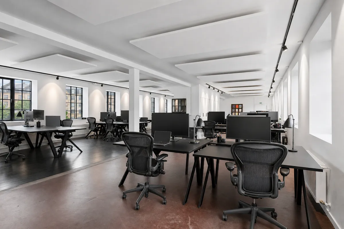 Kontorlokaler med lyse ruder, højt til loftet, og moderne møbler. Sorte borde og skriveborde med monitorer, og ergonomiske stole.