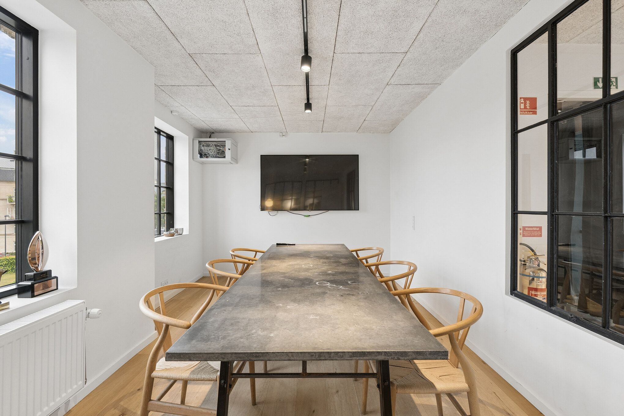 Lys kontor med massiv træbord, lyse og indbydende vinduer, moderne indretning, minimalistiske møbler, og tekniske faciliteter. En tv-skærm er monteret på væggen.