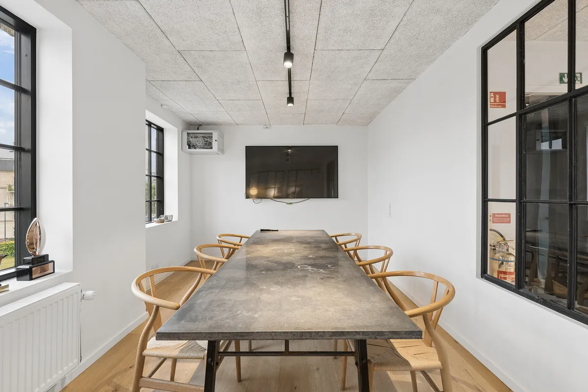 Lys kontor med massiv træbord, lyse og indbydende vinduer, moderne indretning, minimalistiske møbler, og tekniske faciliteter. En tv-skærm er monteret på væggen.