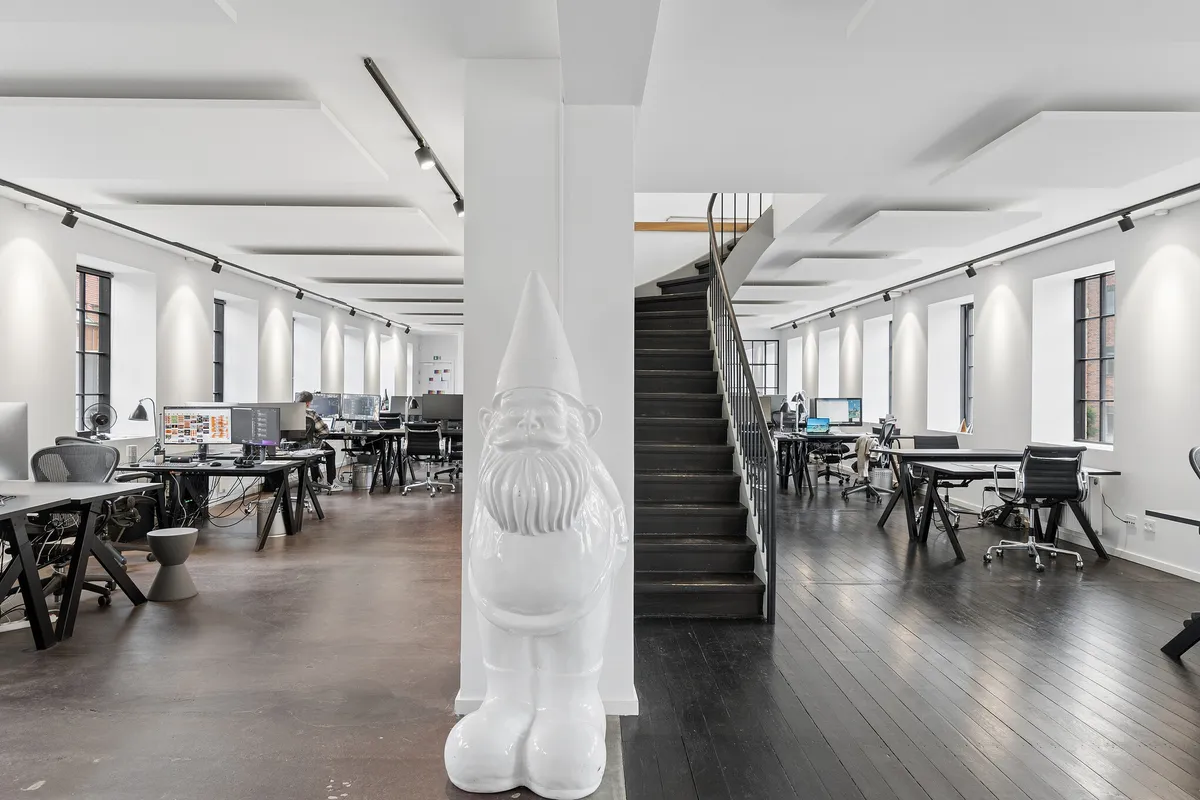 Kontorlokaler med lyse, funktionelle kontorbord, moderne indretning, hvidt loft og gulv, højt loft, kviksølvlampe og loftsvinduer samt en stor, hvid nisse.