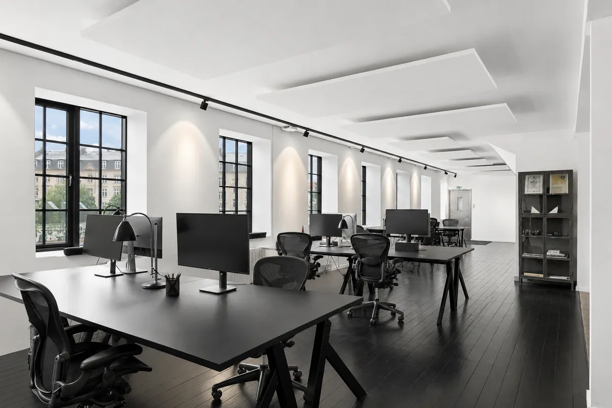 Kontorlokaler med lyse, indbydende rammer og moderne indretning, hvor skriveborde og stolpe udgør et ergonomisk arbejdsområde. Indbygget belysning og store vinduer skaber et produktivt og inspirerende arbejdsmiljø