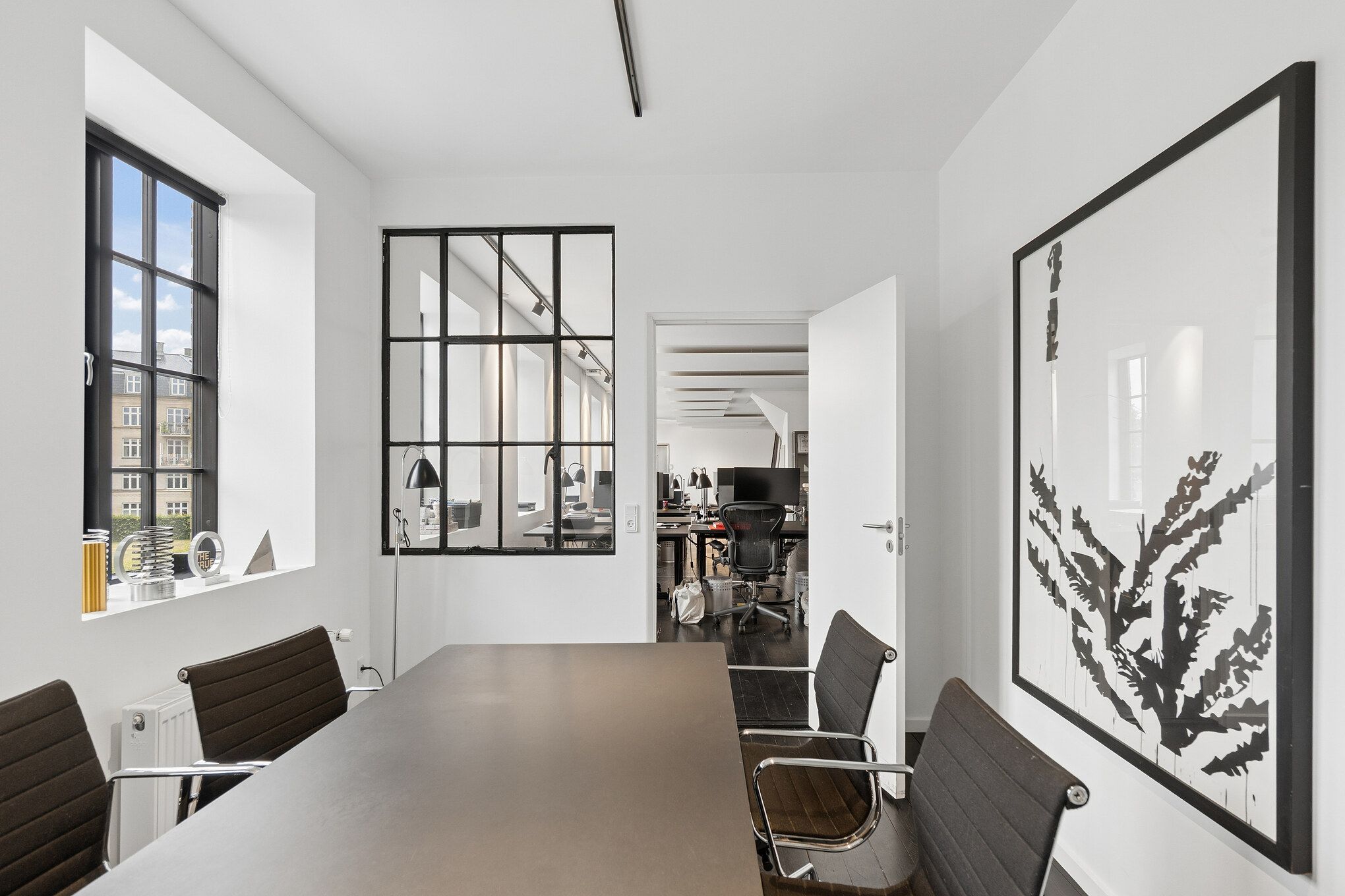 Kontor med moderne møbler, store vinduer, åben dør, lyse vægge og et stort kunstværk med sort og hvid stribet design på væggen