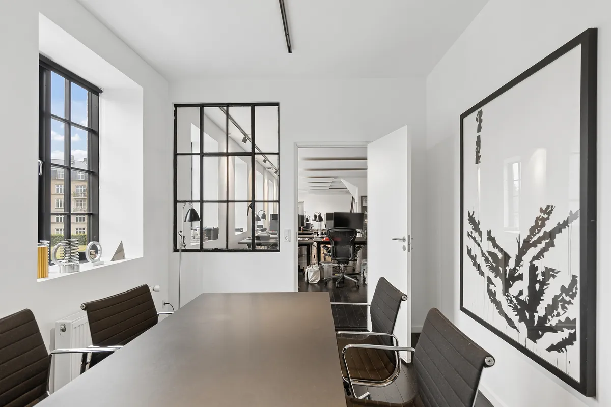 Kontor med moderne møbler, store vinduer, åben dør, lyse vægge og et stort kunstværk med sort og hvid stribet design på væggen