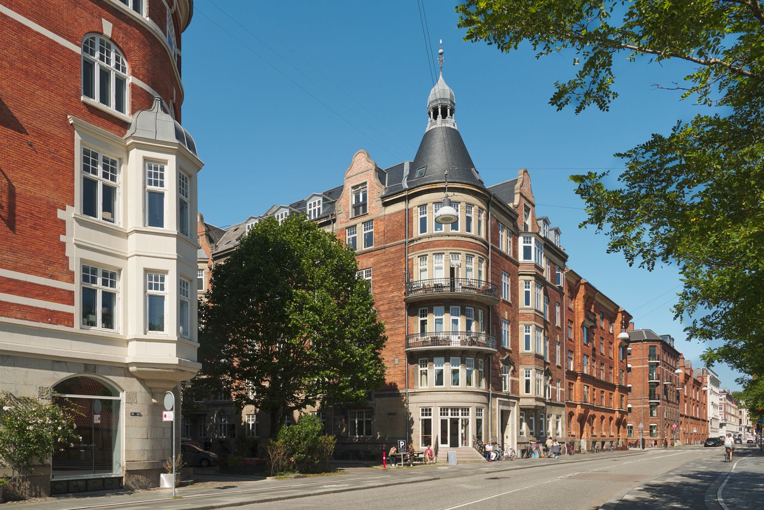 Kontorlejemål med lyse lokaler og store vinduer i en markant hjørneejendom i København. Grønne træer og buske og mennesker på gågaden.