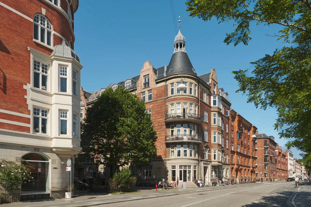 Kontorlejemål med lyse lokaler og store vinduer i en markant hjørneejendom i København. Grønne træer og buske og mennesker på gågaden.