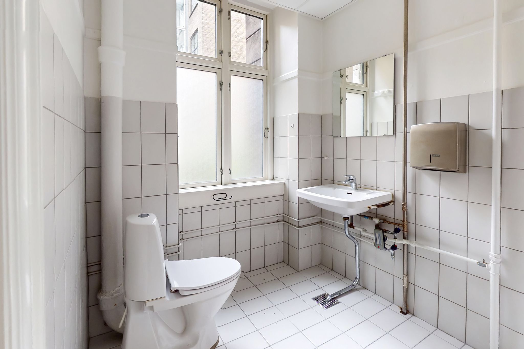 En hvidt klædt badeværelse med toilet, håndvask og spejl samt en håndklædevarmer og ventilationsrør på væggen