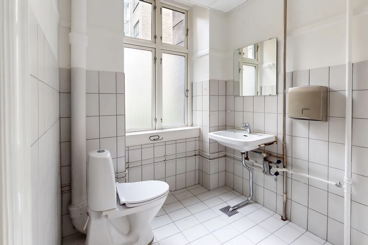 En hvidt klædt badeværelse med toilet, håndvask og spejl samt en håndklædevarmer og ventilationsrør på væggen