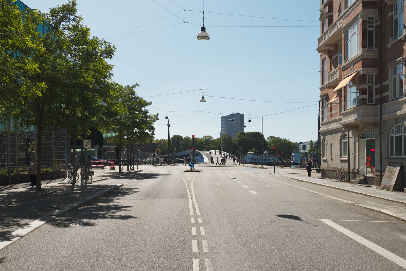 Bred gade med smal asfaltbelægning og huse. Gågade og stier med mennesker og cykler. Høje bygninger og busser i baggrunden. Søjler og elektriske ledninger over gaden.