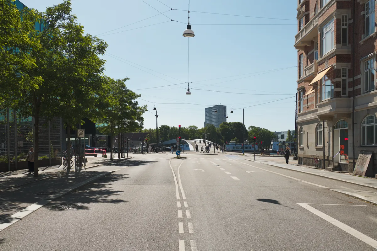 Bred gade med smal asfaltbelægning og huse. Gågade og stier med mennesker og cykler. Høje bygninger og busser i baggrunden. Søjler og elektriske ledninger over gaden.