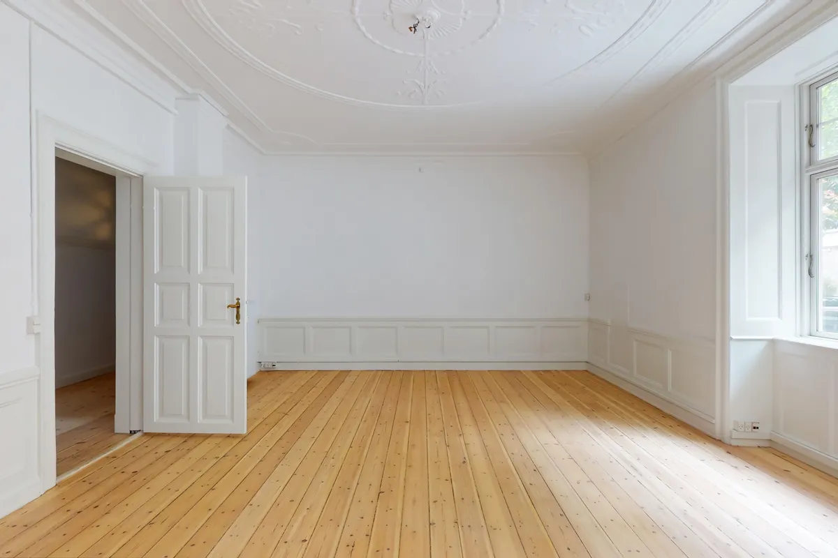 Rummelig herskabelig lejlighed med højloft og trægulv, to døre, hvidmalede vægge og lyse vinduer med solskin