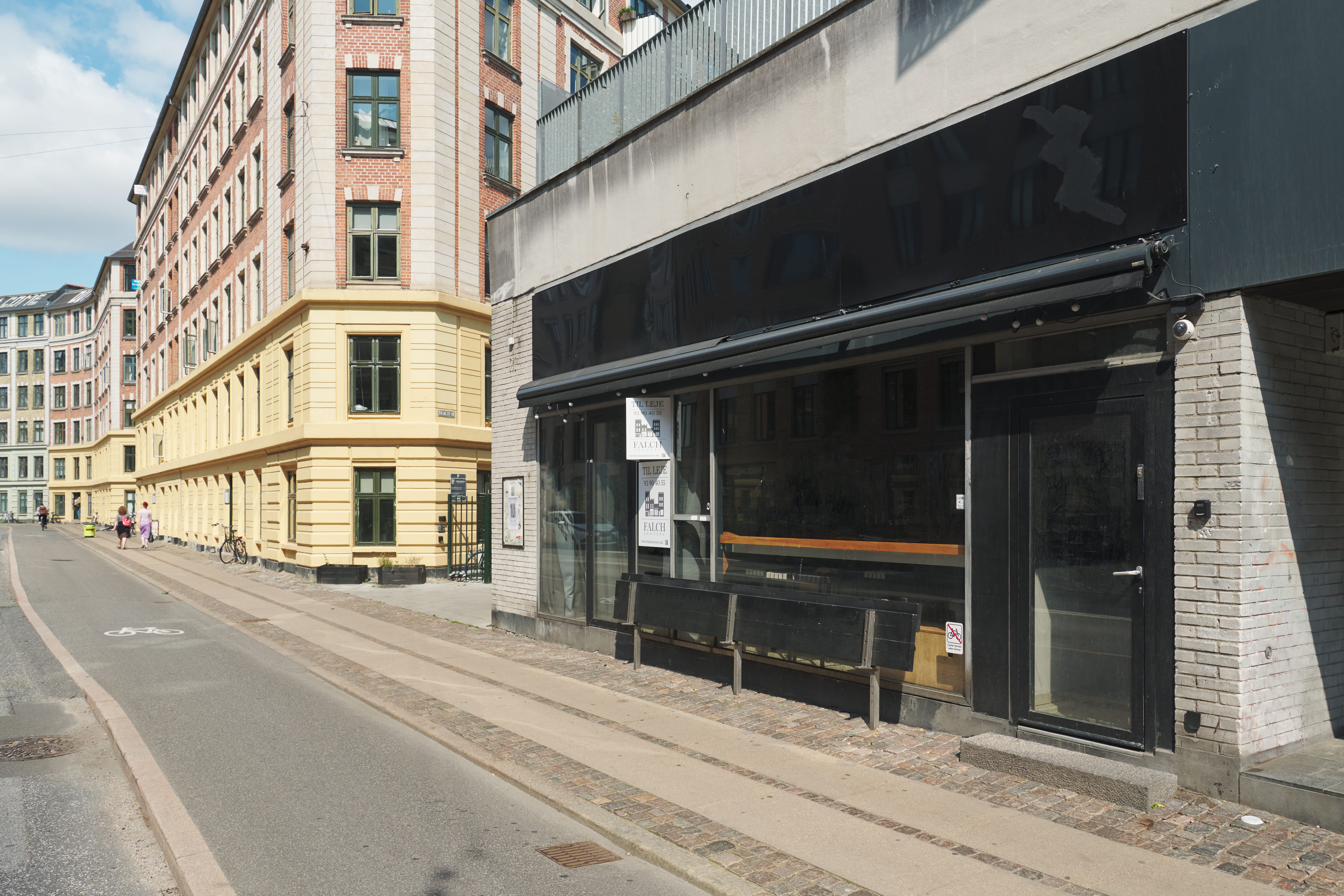Strøget med to huse og café. En butik med mørkt glas og tegning på facaden. Foran er to bænke og skråkanter. To personer og to cykler i baggrunden.
