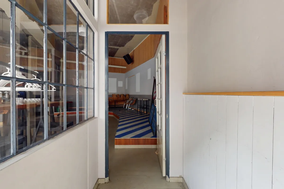 En lang hal med lyseglas og hvid væg. I enden af hallen er der en dør til en stue med møbler og gulvbelægning i blå og hvide striber