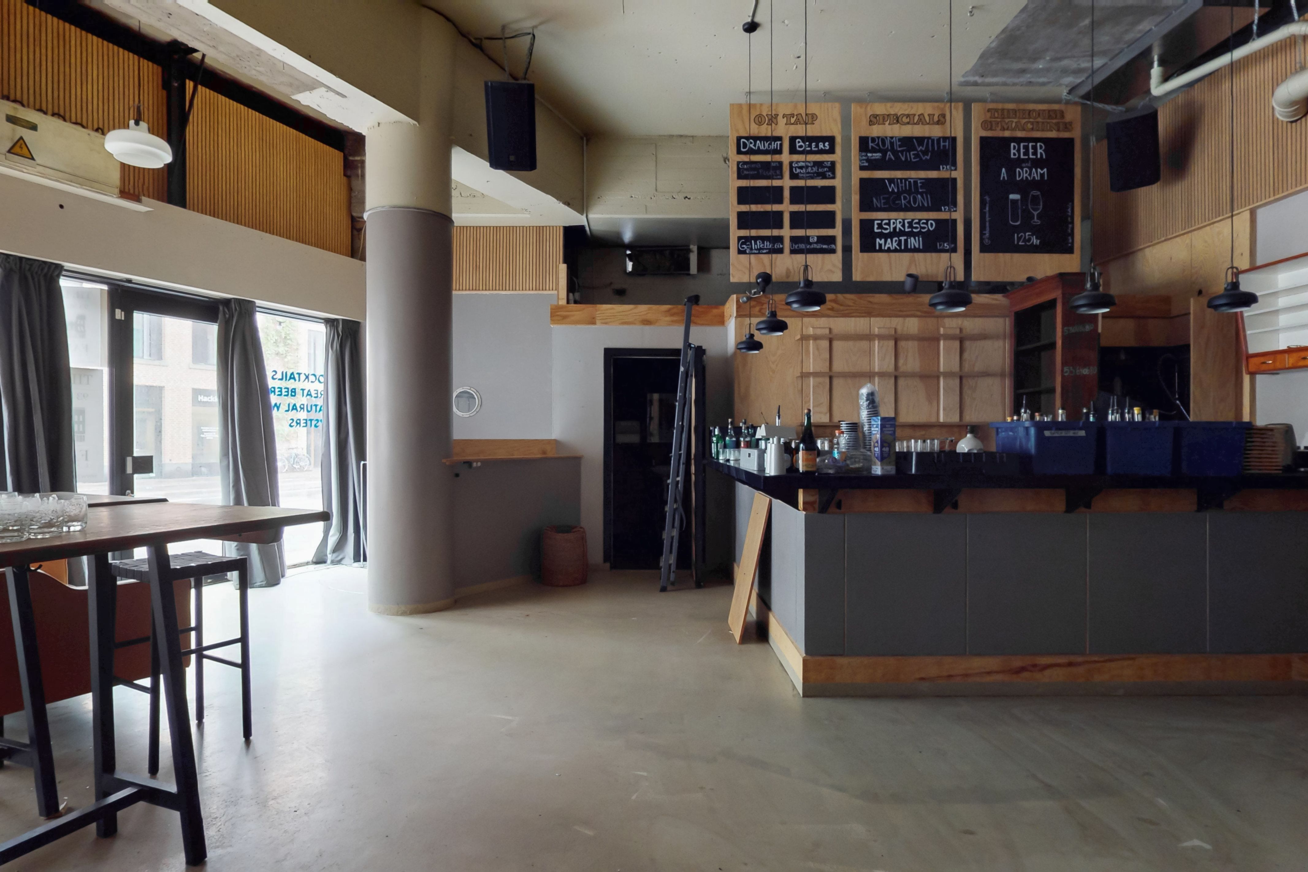 Cafe med trægulve, loftsbjælke og højt loft. Sort køkkenbord med flasker, glas og øl. Sorte lysekopper og lysekroner. Menubræt med øl, drikkevarer og specialiteter.