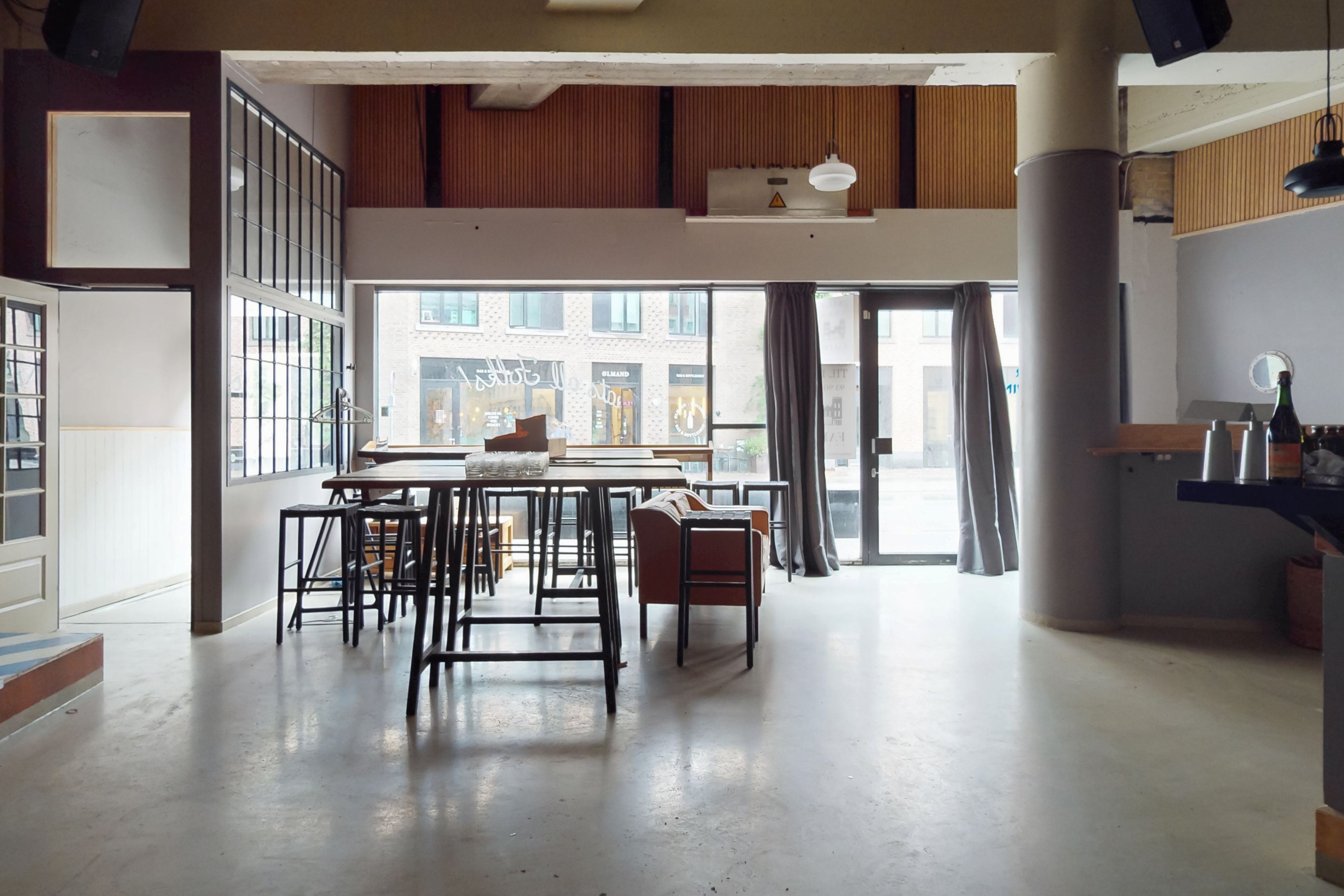 Moderne café med store vinduer, møbler og træpaneler, en flaske vin og en kuffert på et stolpebord og et skab med flere flasker og glas