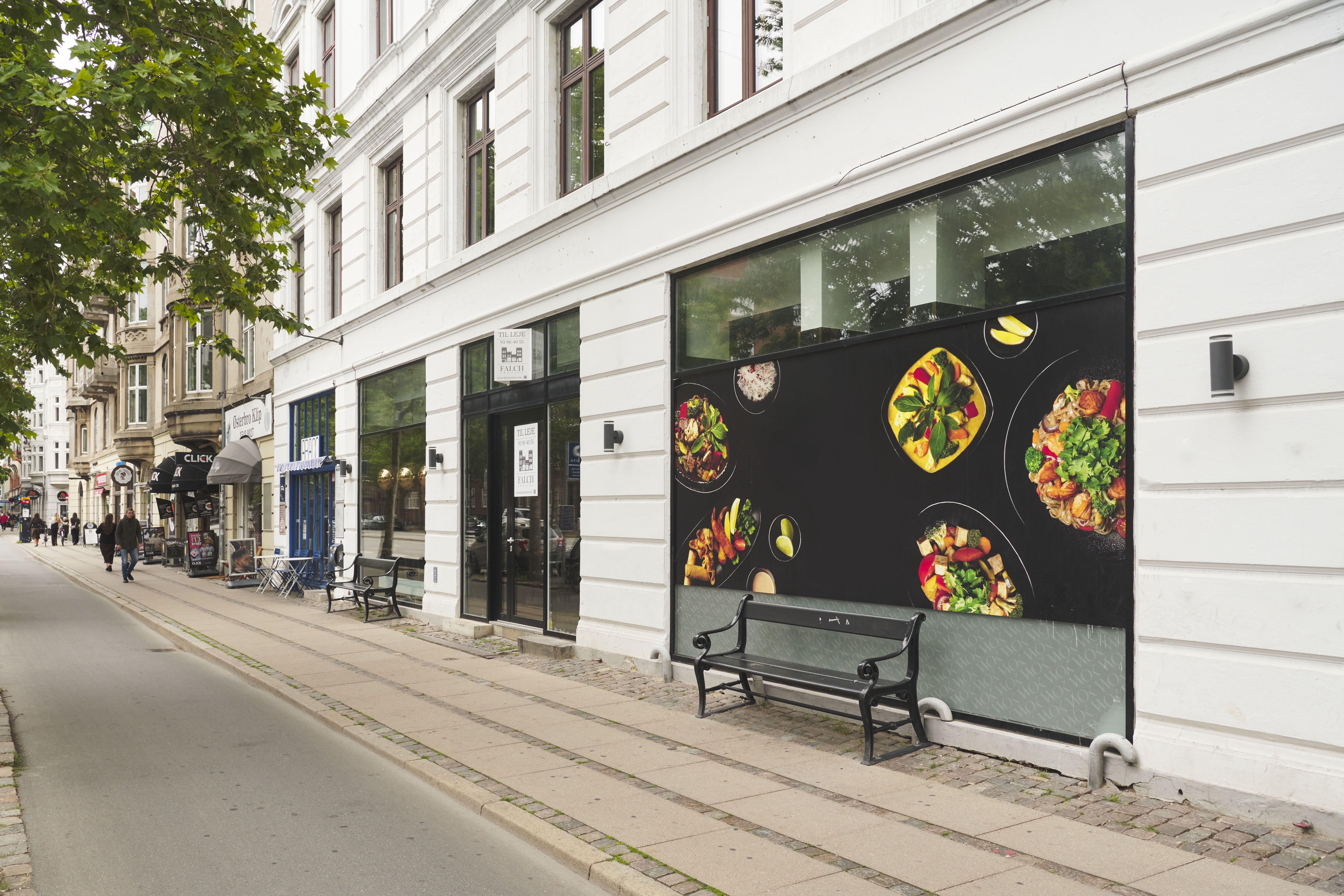 Større og mindre stole og borde udenfor restauranten. En person går langs fortovet og går ind i en butik. En sort sofabank på fortovet. Et maleri af en stor tallerken med føde på facaden.