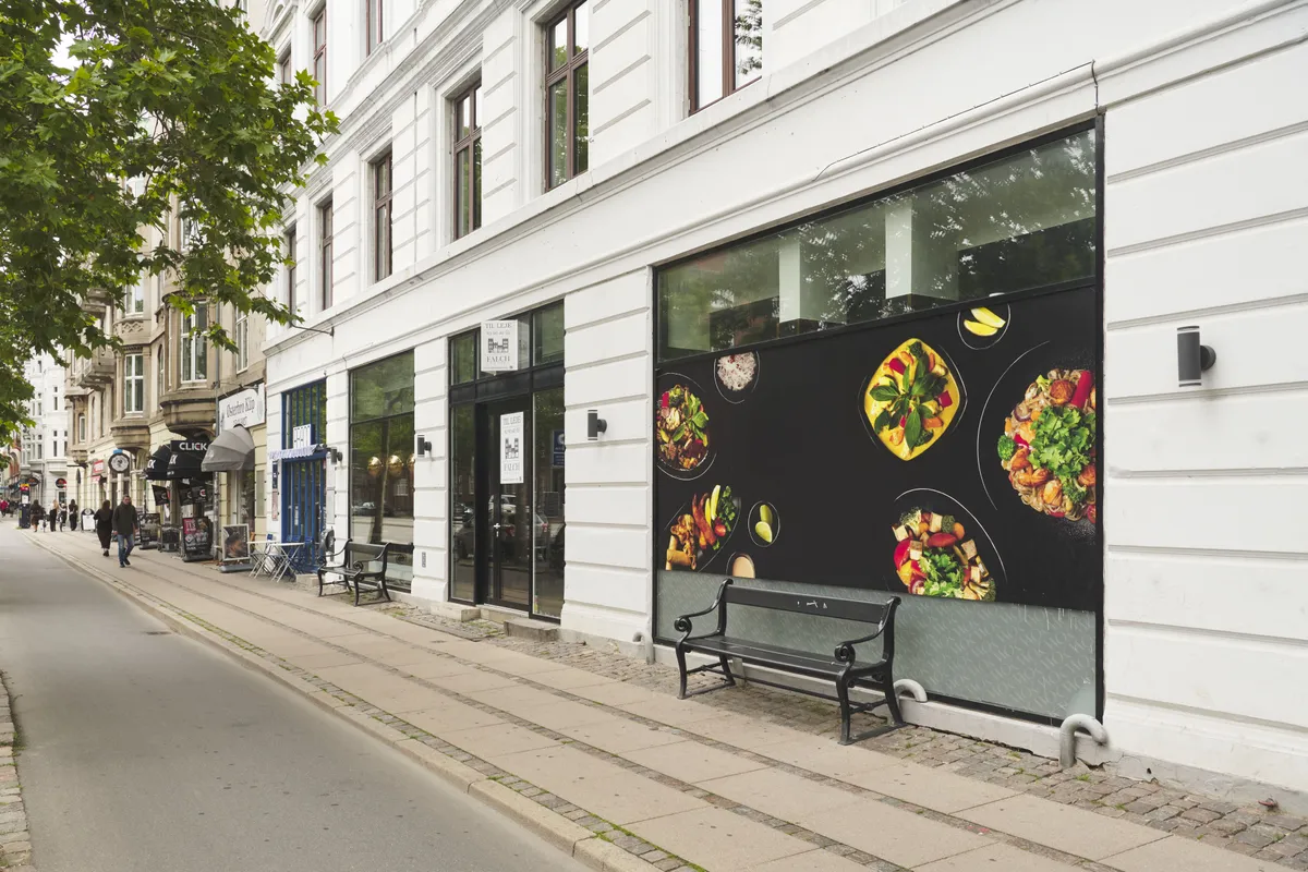 Større og mindre stole og borde udenfor restauranten. En person går langs fortovet og går ind i en butik. En sort sofabank på fortovet. Et maleri af en stor tallerken med føde på facaden.