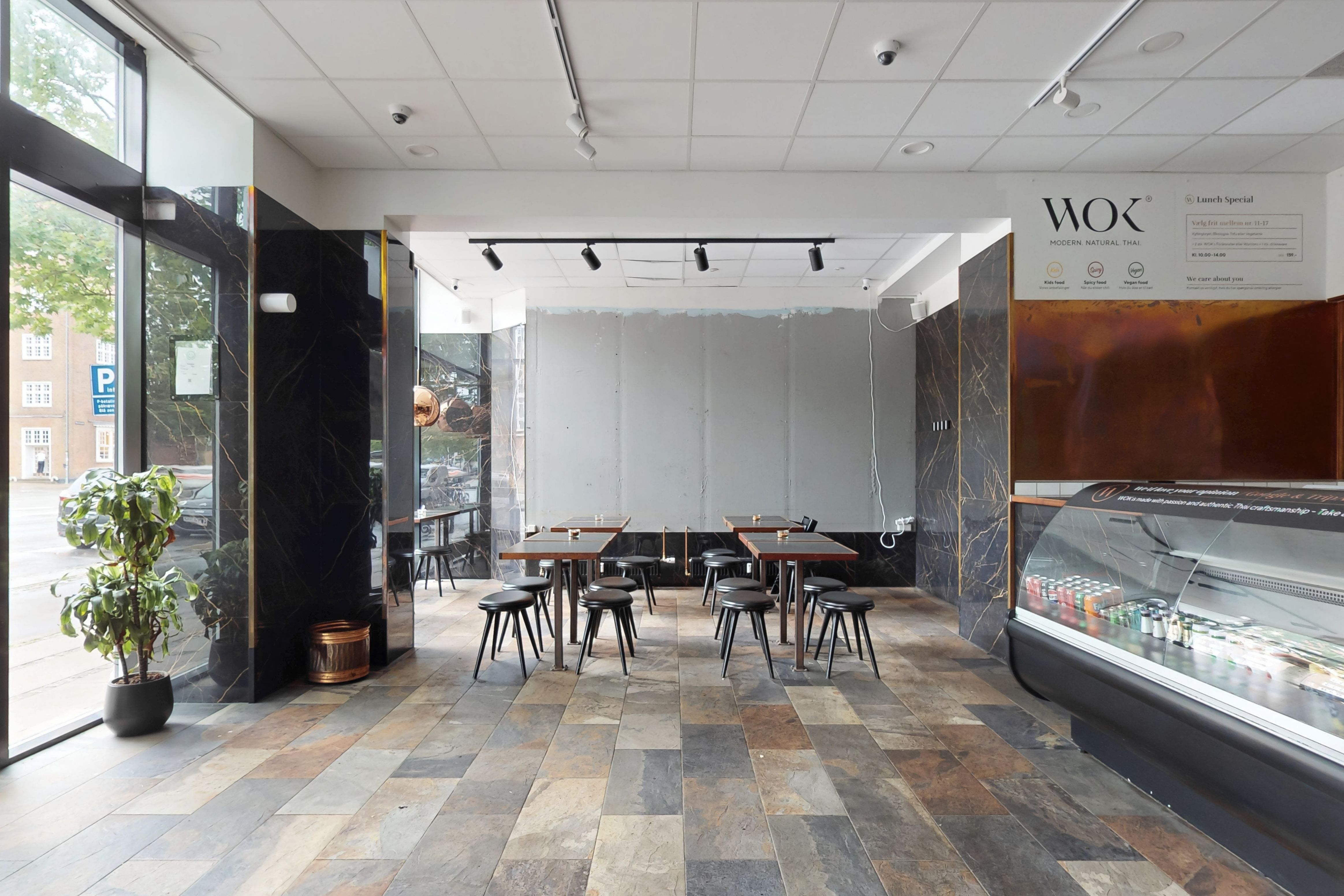 Indvendig i en restaurant med trægulv og moderne møbler samt udsigt til gade og park. Der er en glasvitrine med drikkevarer og en menuvæg med logoet for Wok Modern Natural Thai