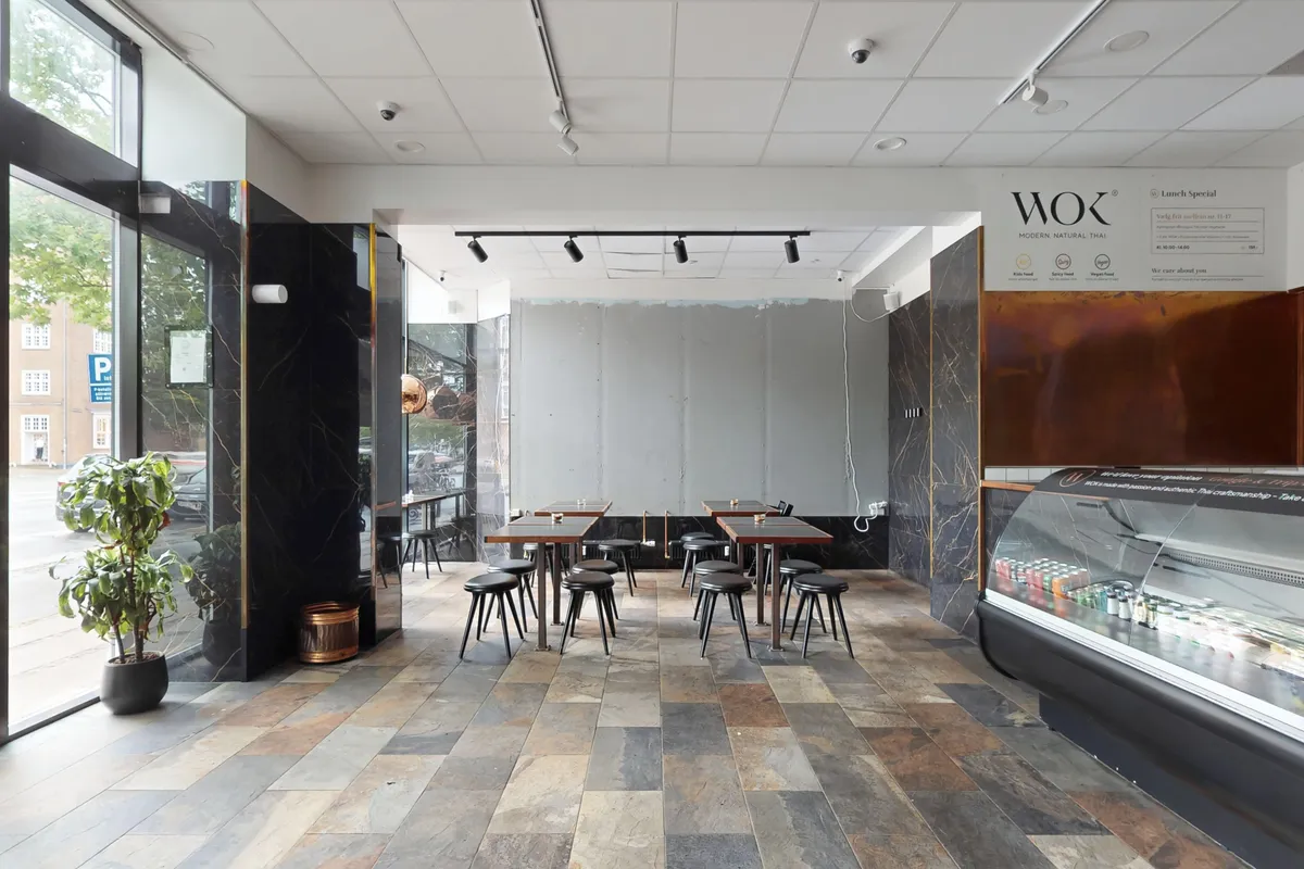 Indvendig i en restaurant med trægulv og moderne møbler samt udsigt til gade og park. Der er en glasvitrine med drikkevarer og en menuvæg med logoet for Wok Modern Natural Thai