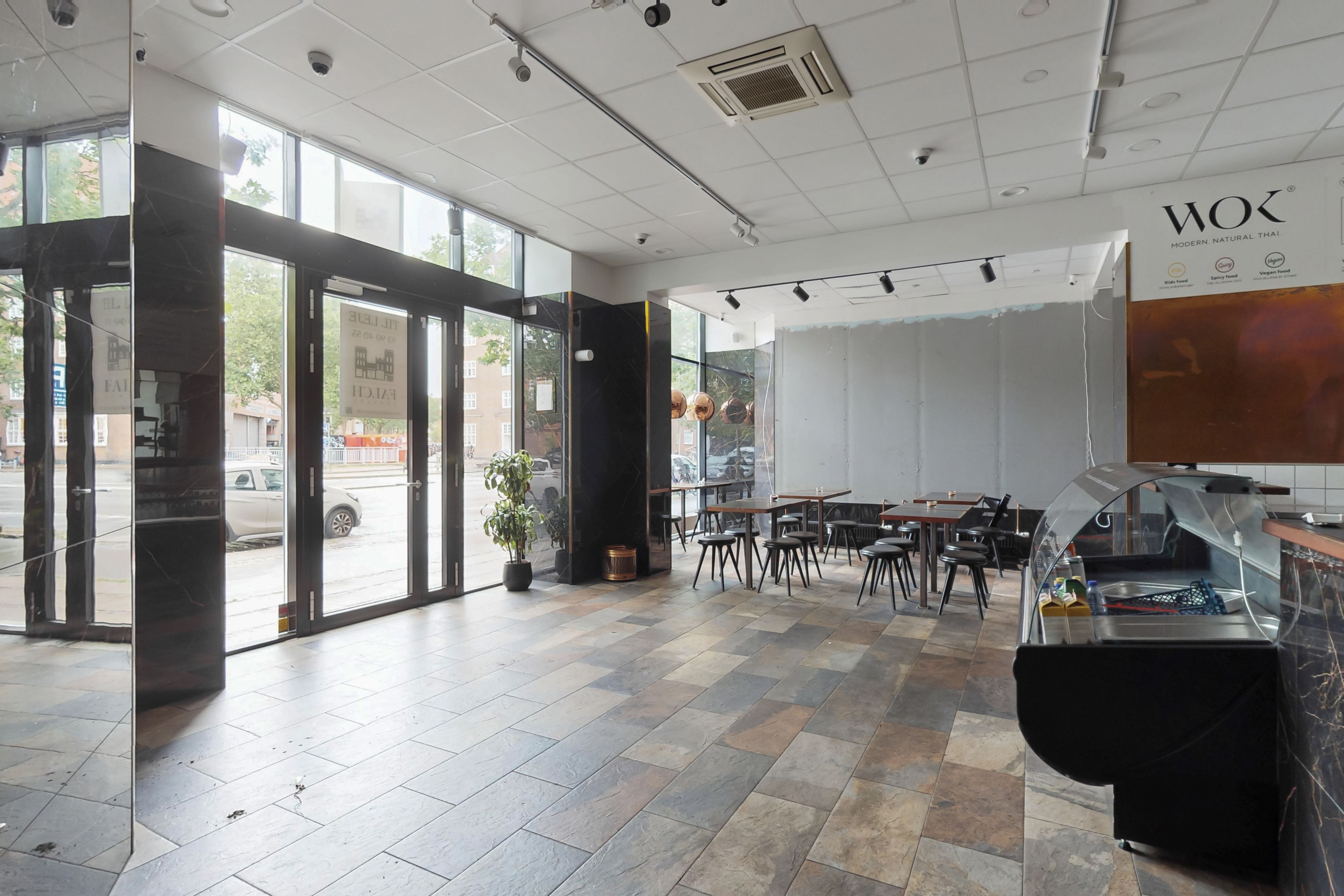 Inde i restauranten med moderne naturlige thailandske retter er der to store vinduer og et åbent gulv med krydsfinerbelagt træ. Der er to små borde med stole.