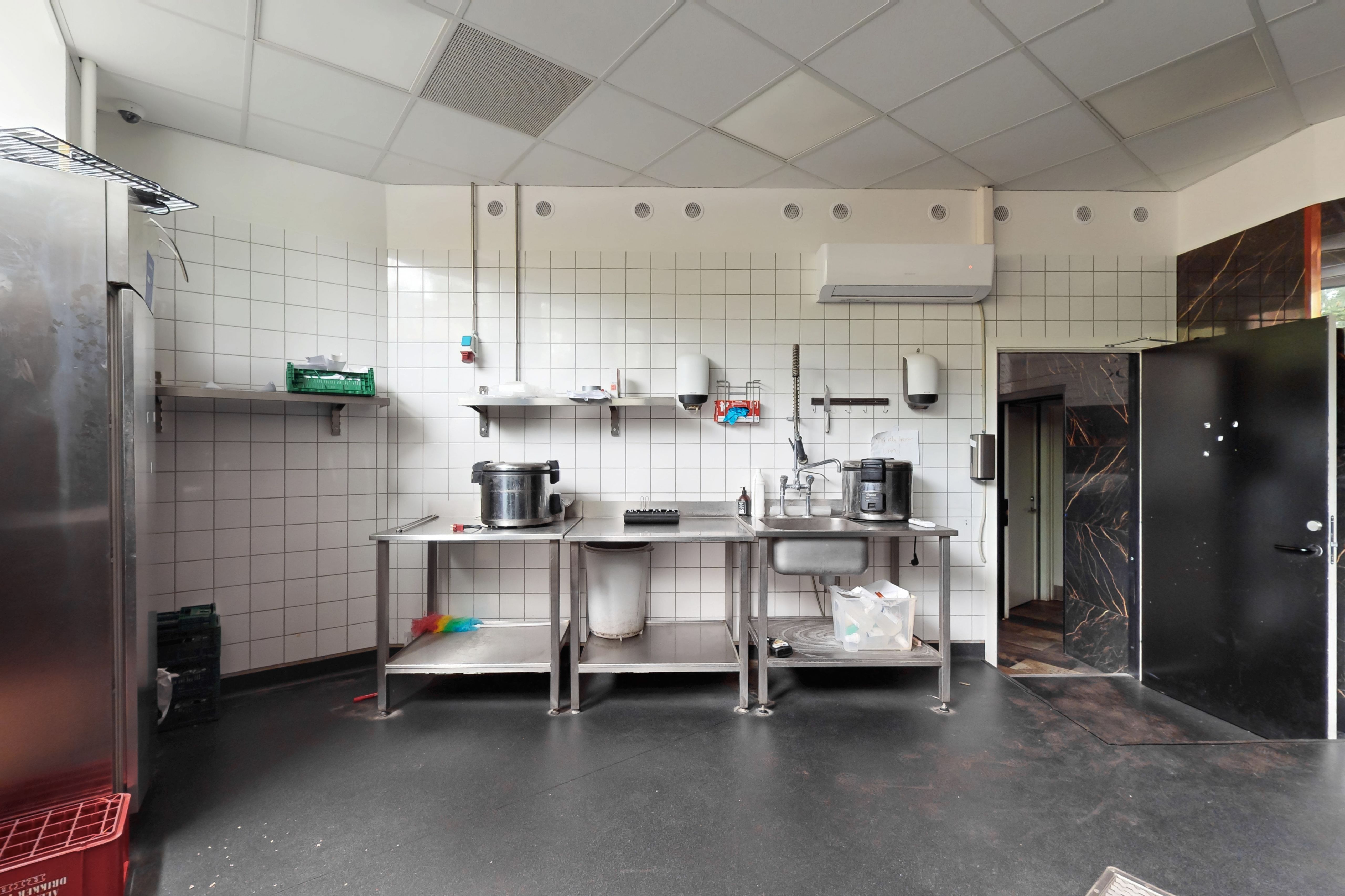 Stor køkken med flere stålborde, fryseskab og rødt opbevaringskurv samt åbne døre til siderum. Køkkenvæggene er tæt beklædt med hvide fliser og udstyret med flere væghylder og håndvask