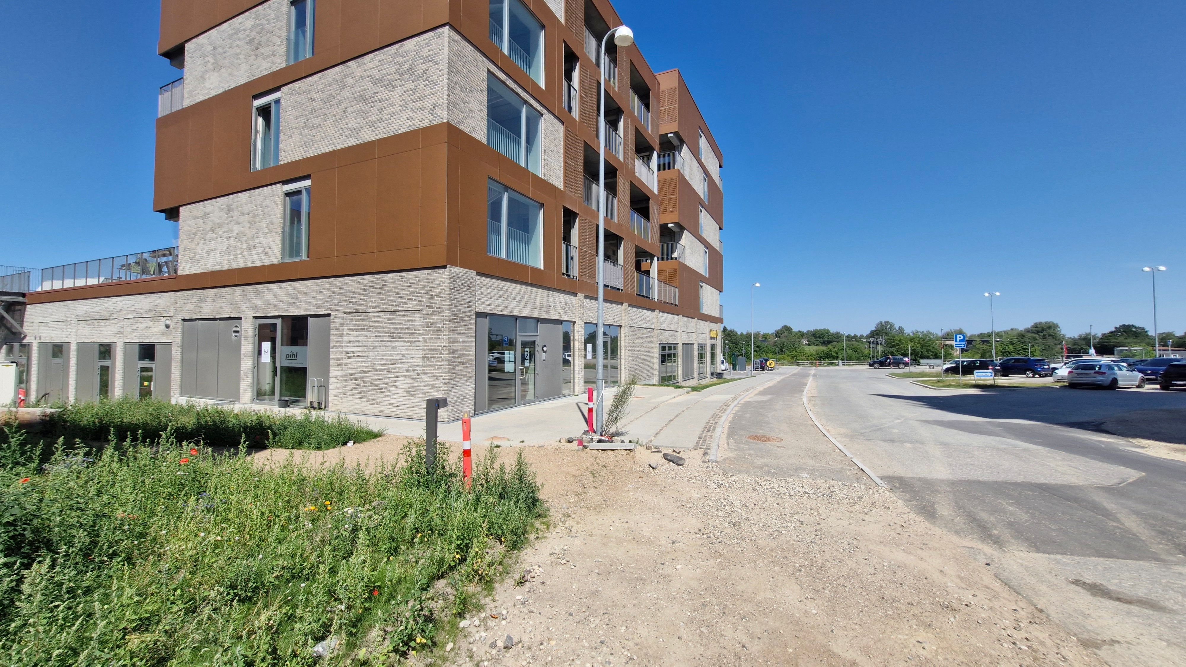 Lejlighedsbygning med moderne design og store ruder. Adgangsvej og parkeringsplads. Solrige og fredelige omgivelser med grønne planter og buske.