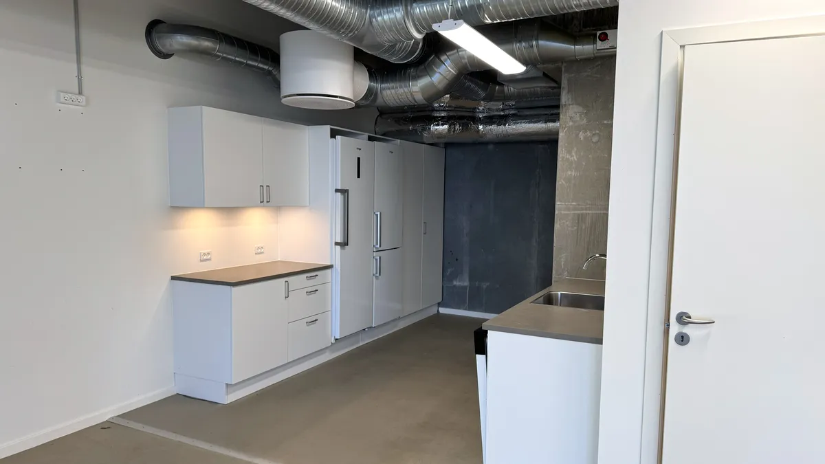 Moderne køkken med hvide vægge og lyse lofter. Køkken med lyse, hvide skabe, lysegråt bordplade og indbygget køleskab. Døren til køkkenet er åben, og der er en rustfri stålvæskedispenser monteret på væggen.