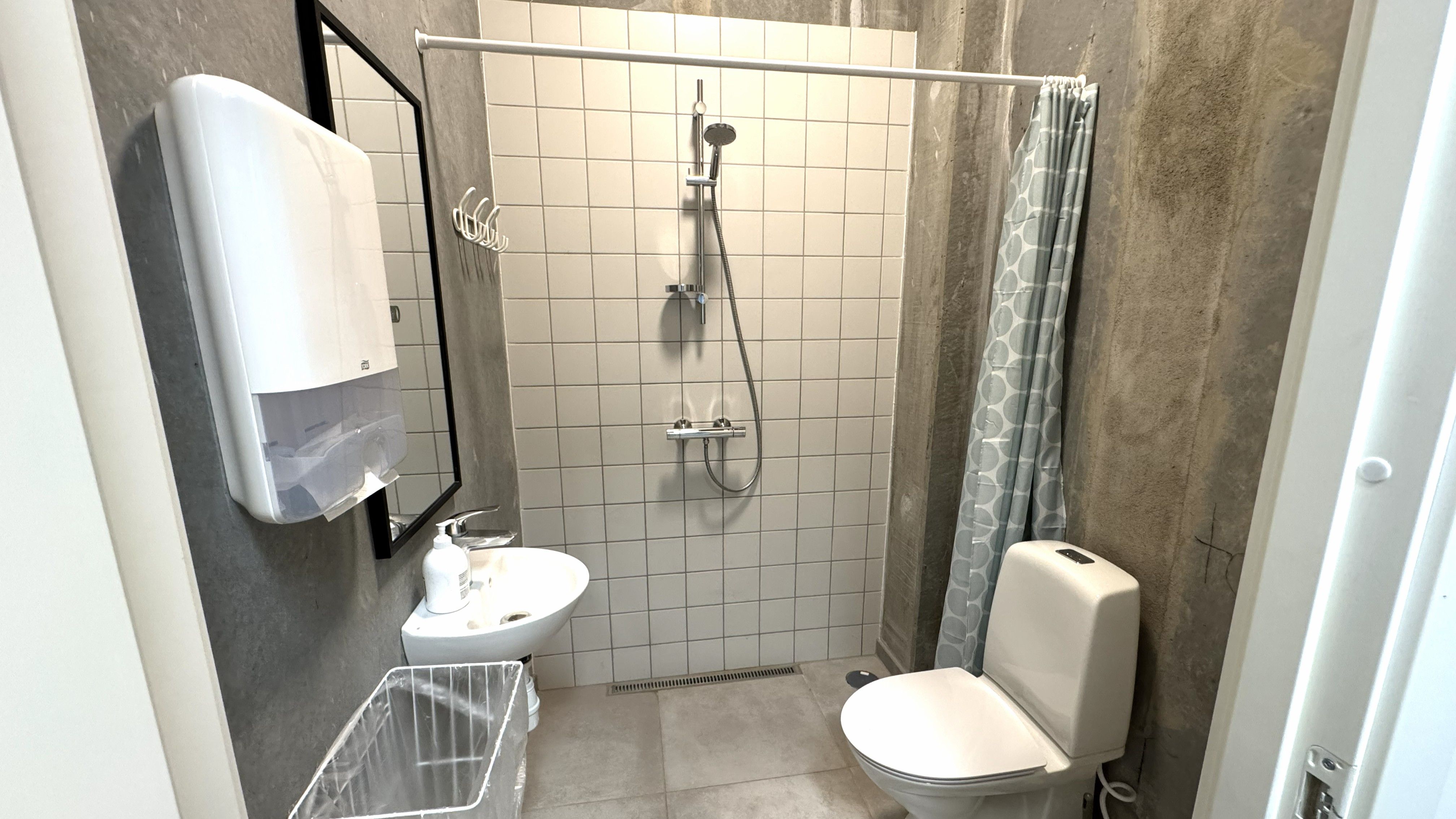 Et moderne badeværelse med hvidt keramik og hvidt håndklæde. Håndklæde er hængende i et stålhylde. Et hvidt toilet er udenfor brusebadet.