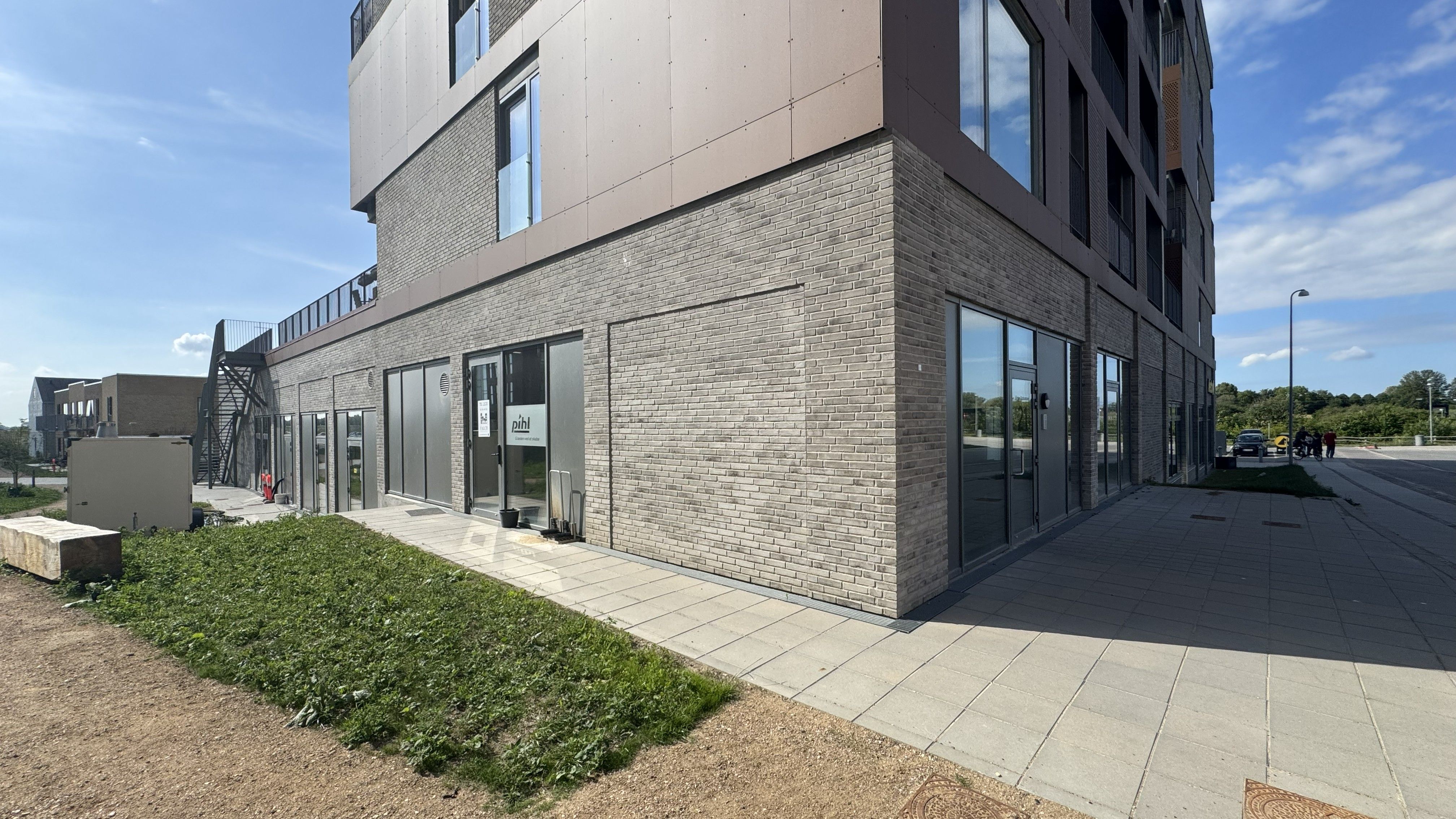 Moderne erhvervslejemål med glasdøre og store vinduer på Vinge Stationsvej 23. Lys gulv og grønne anlæg. Solrig dag med lyse skyer i baggrunden.