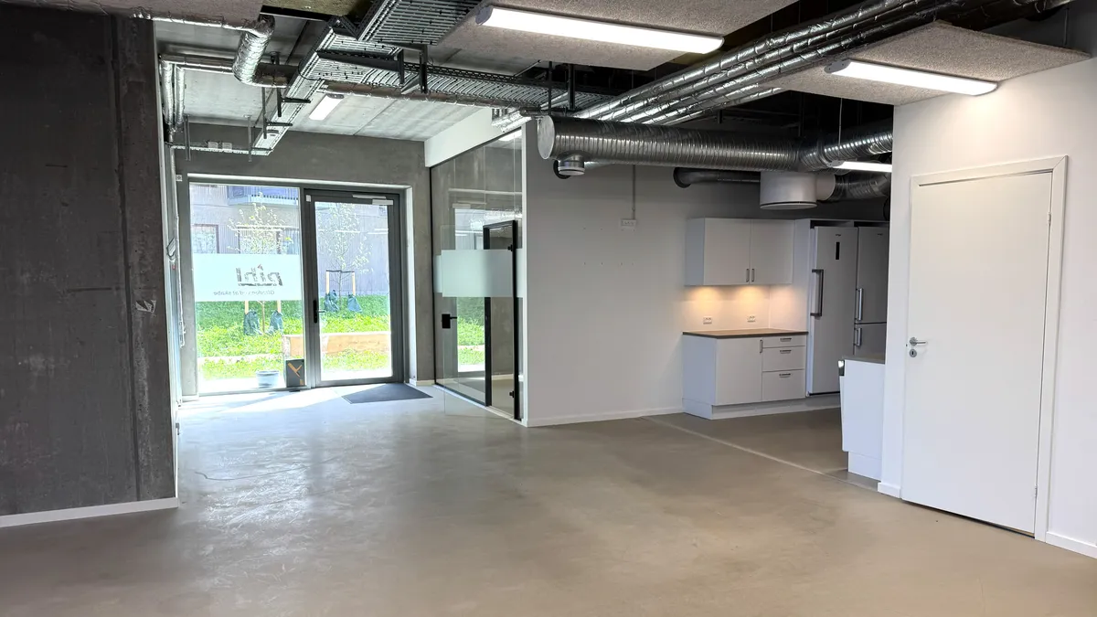 Moderne kontorrum med glasdøre, lyse loftslampe, hvide vægge og trægulv. Der er køkken med hvide skabe og høj køleskab. Der er et rengøringsrum med en opvaskeskur. Der er reklameskilt og bygningsstrukturer udenfor.