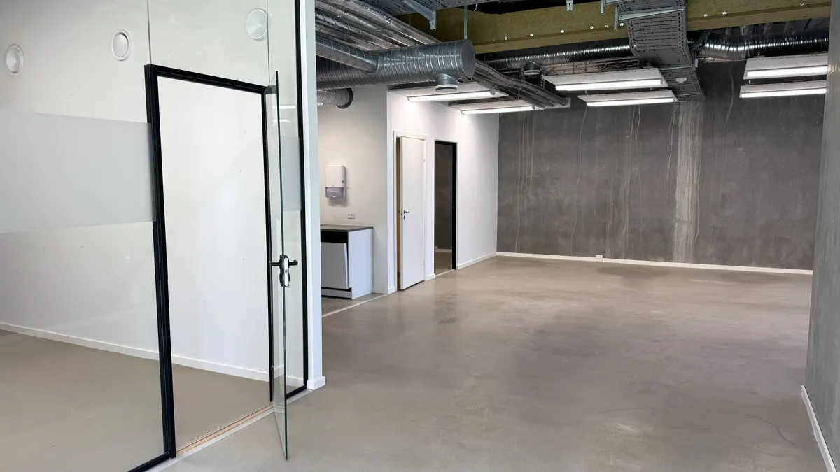 En stor og lys kontordør med glas og sort ramme. Et moderne kontorlokale med hvid og grå væg. Store lyse buer og hvide loftslampe. En hvid børste og en hvid toilet.