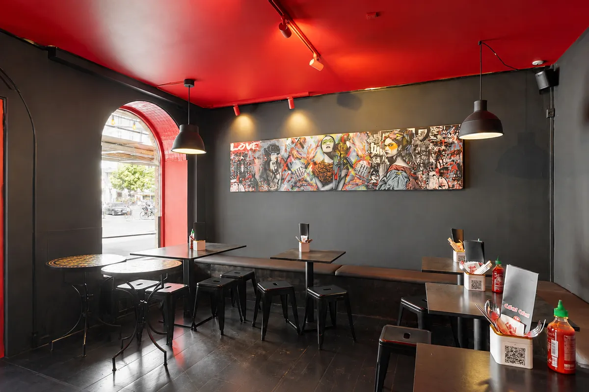 En elegant café med sorte og røde vægge, et stort maleri, runde borde og stole, og lysekroner. Et rødt glasvindue udenfor viser en vej med biler