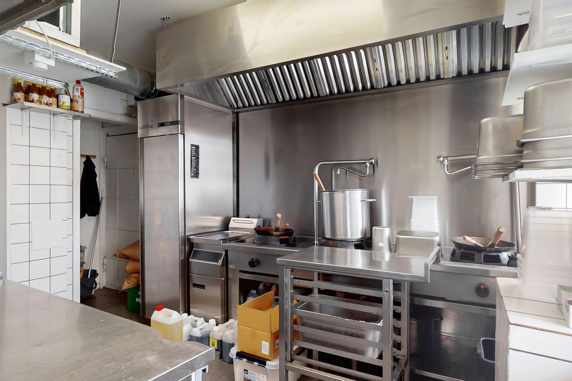 En nydelig café med moderne køkken og apparater, spisesteder og et højt loft med ventilationsblæser. Flasker og bøtter står klar til brug, og der er en dør til et køleskab