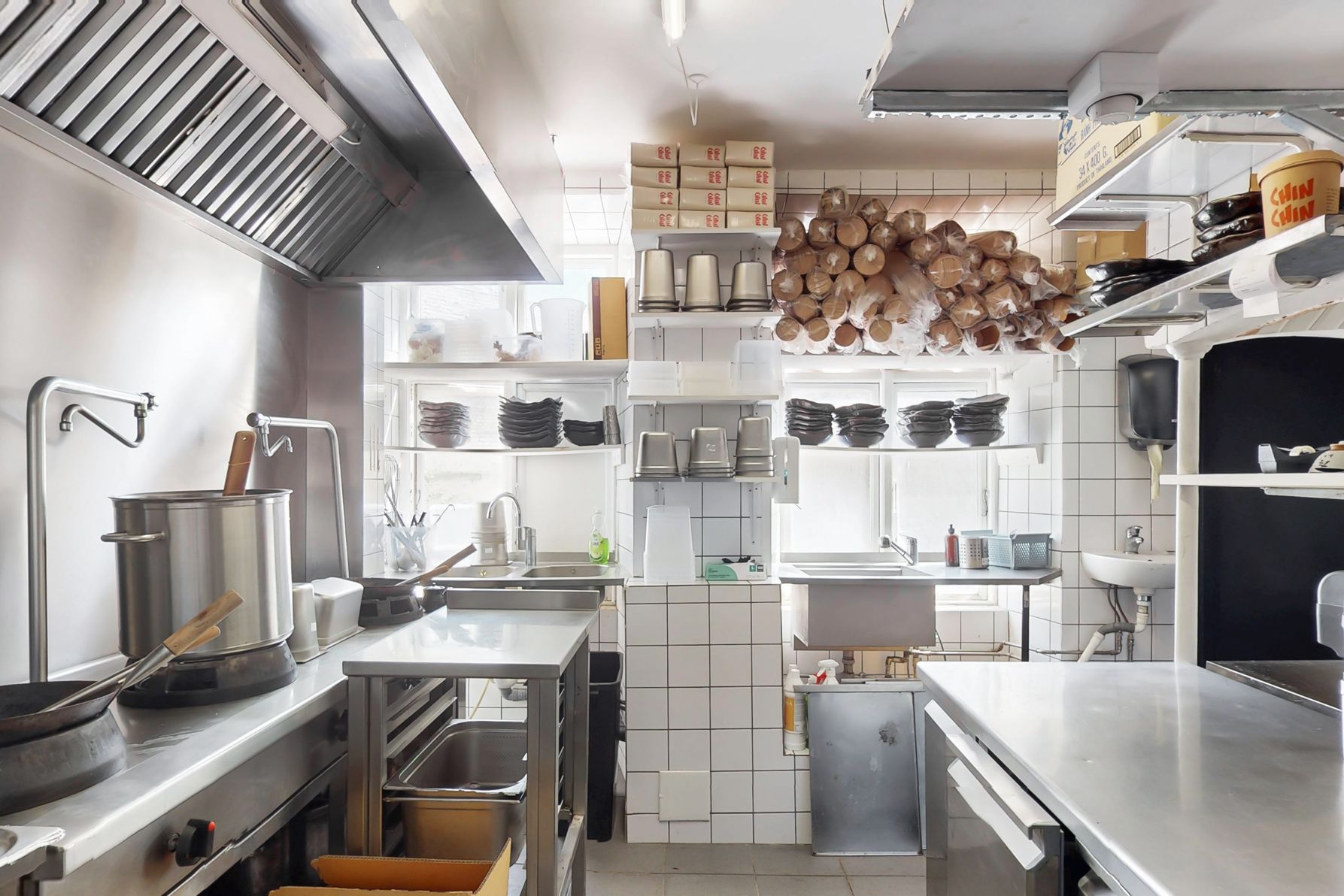 En ren café med højt til loftet, flere ståloverflader, kedler, gryder, og en stor køkkenudstyr. Fladt, brændenælde-frit gulv og store vinduer til lys.