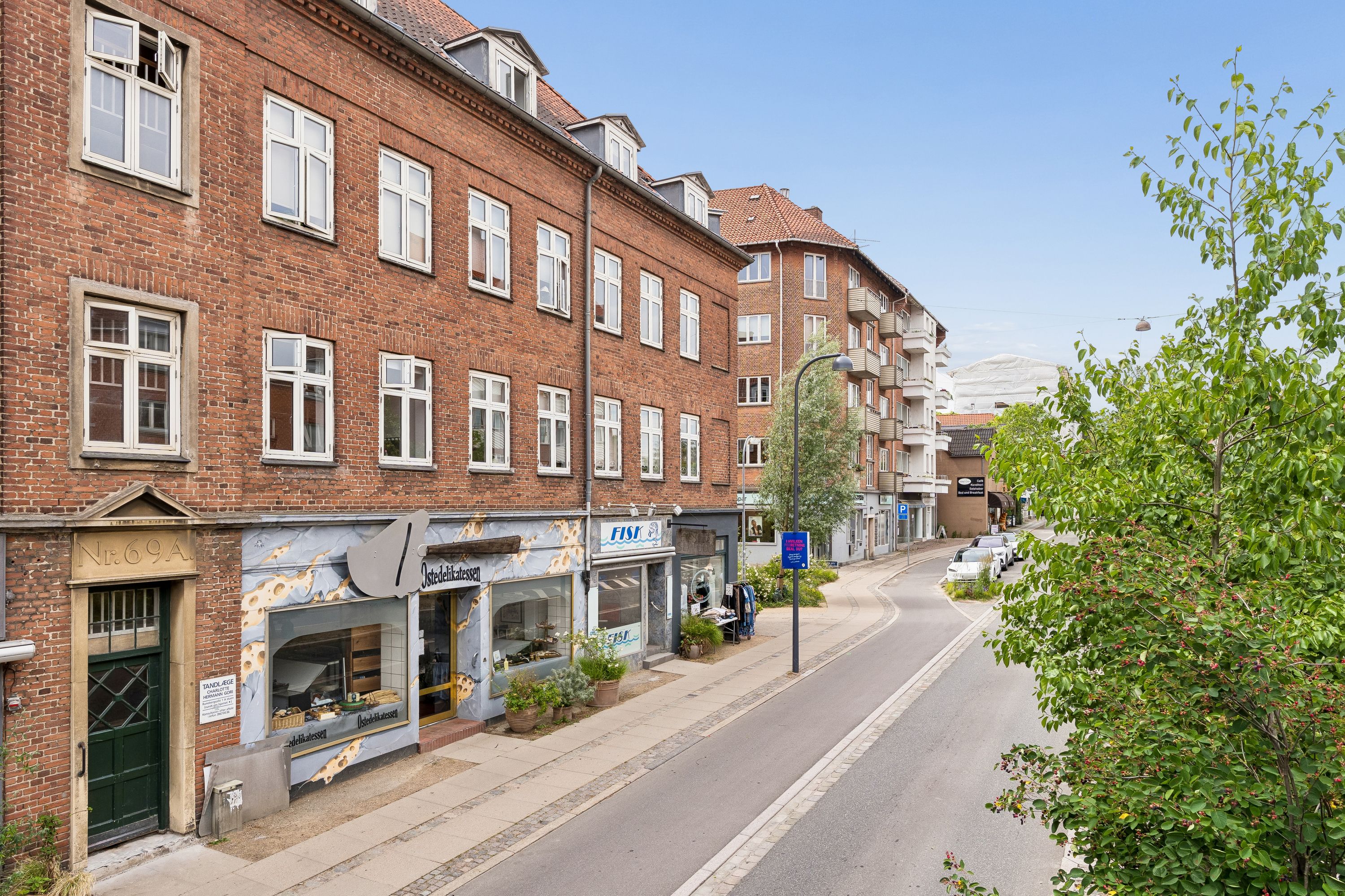 Butik til leje på Søborg Hovedgade 69A, st. 8. Butikkerne har store ruder og er lavet af røde teglsten. Der er træer og buske langs fortovet