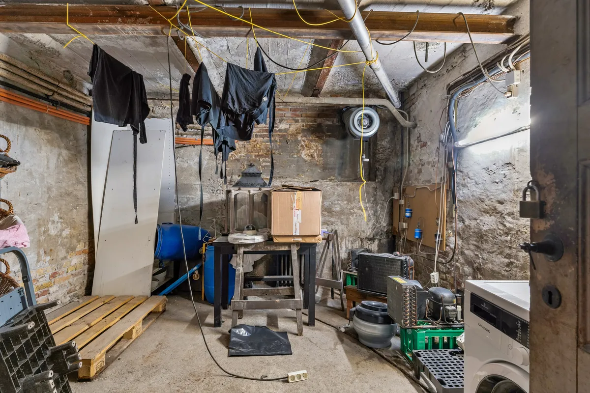 En lysegrå tøjstativ med to tørrede tøjstykker hængende. En stor kasse og en vaskemaskine står på gulvet. Der er et ventiler og elektriske ledninger over gulvet.