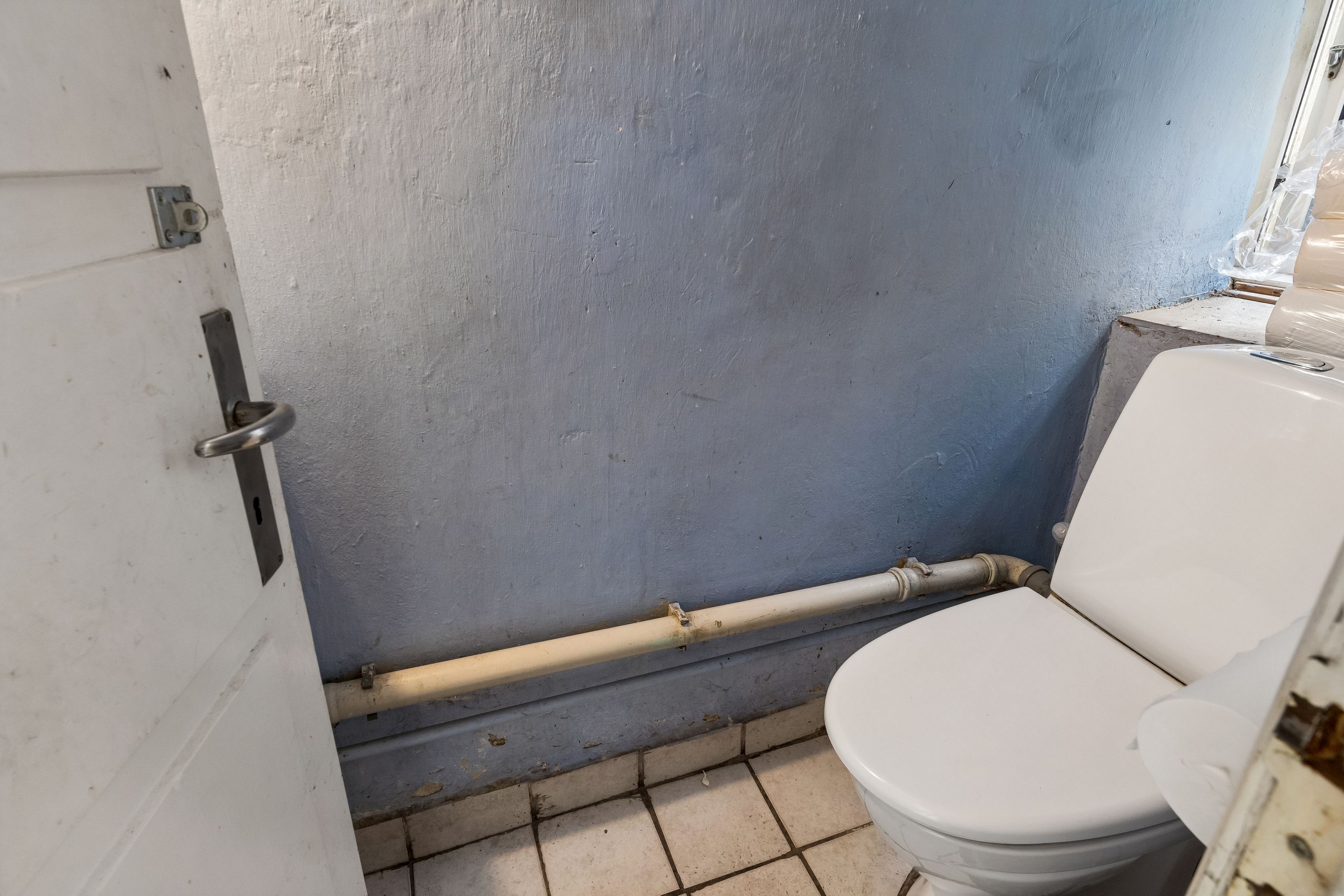 En gammel toilet i et nedslidt rum med hvide vægge, en toiletblok og rør tilsluttet til væggen og en hvid dør med håndtag og lås