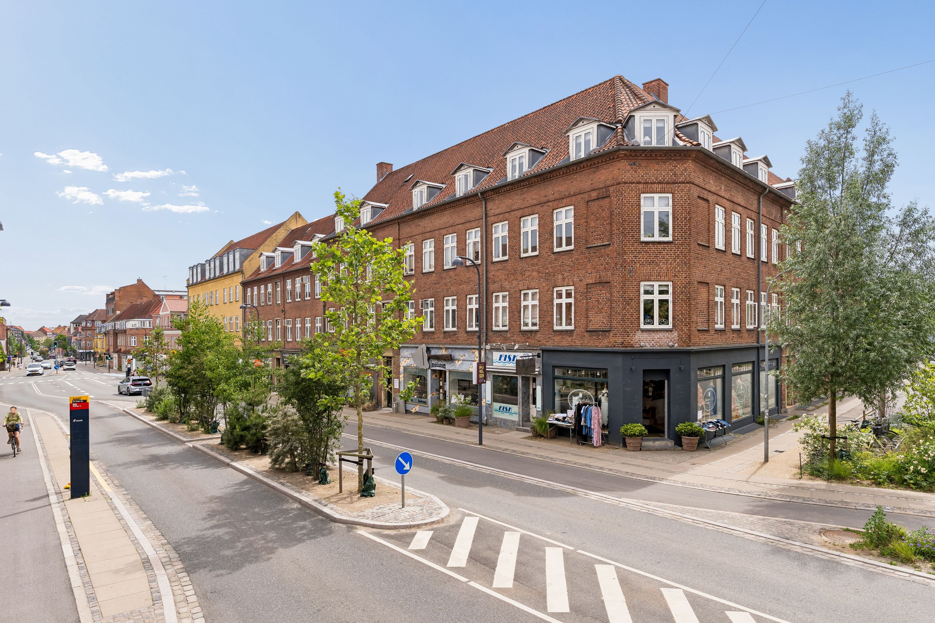 En cykelsti og to butikker med FISS-skilt. Der er et nyt cykelfortov og grønne anlæg ved siden af. Vejen er tom og der er kun en cyklist på vejen