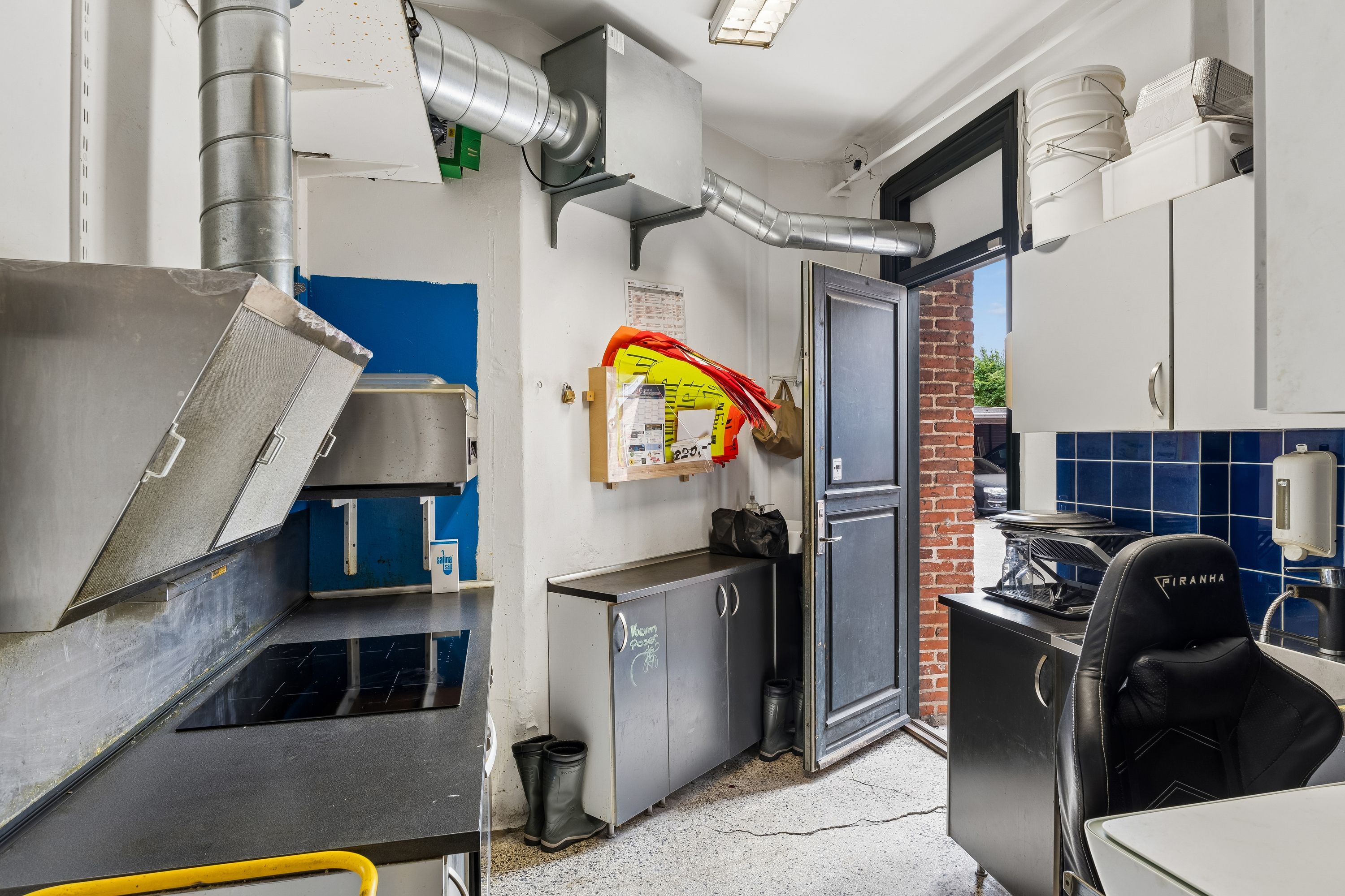 En forretningslokal til leje med køkken med højt loft, metal apparater, loftsmonteret ventilation og opbevaringsskabe. Der er åben dør til udendørsområdet og en fælles opvaskemaskine.