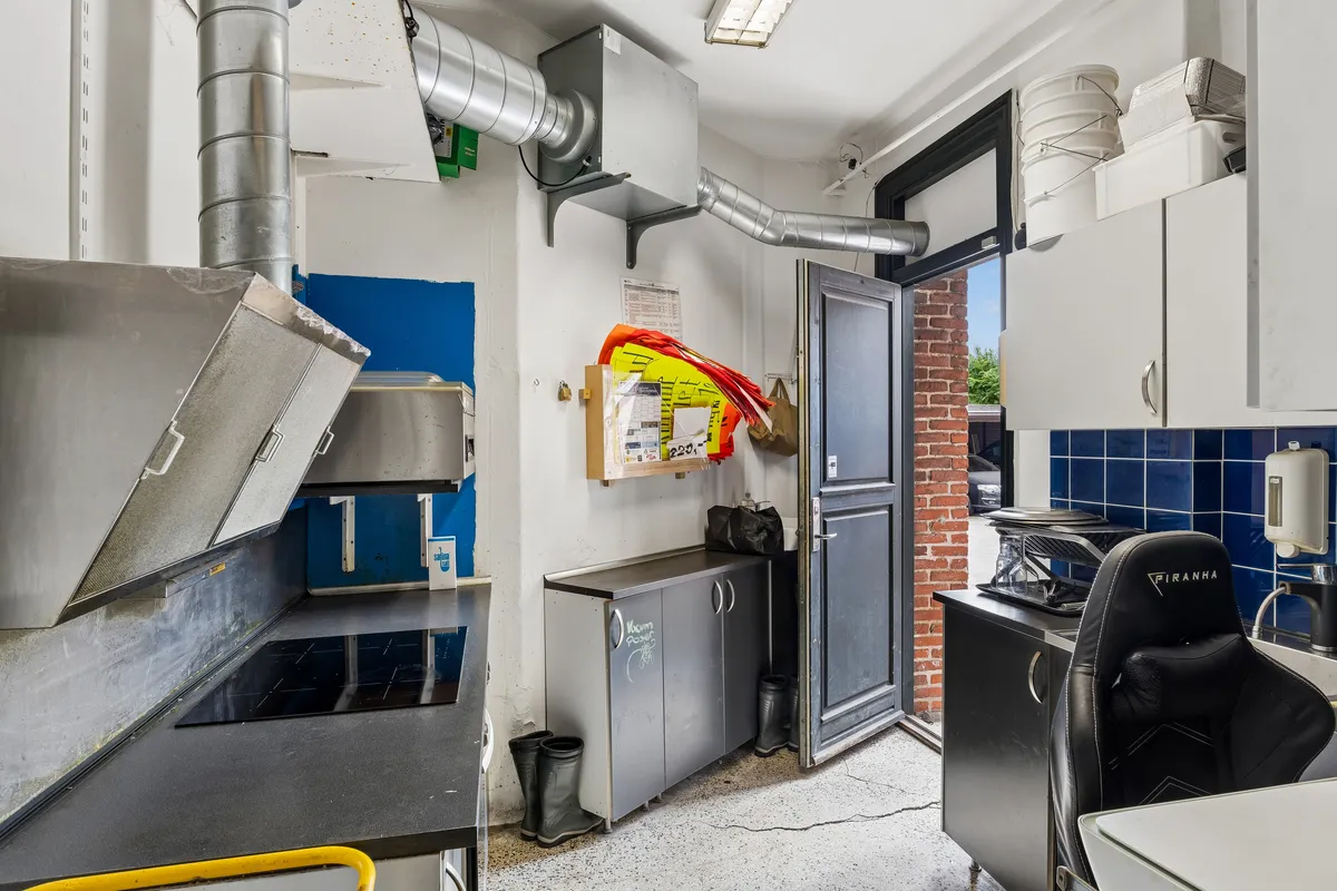 En forretningslokal til leje med køkken med højt loft, metal apparater, loftsmonteret ventilation og opbevaringsskabe. Der er åben dør til udendørsområdet og en fælles opvaskemaskine.