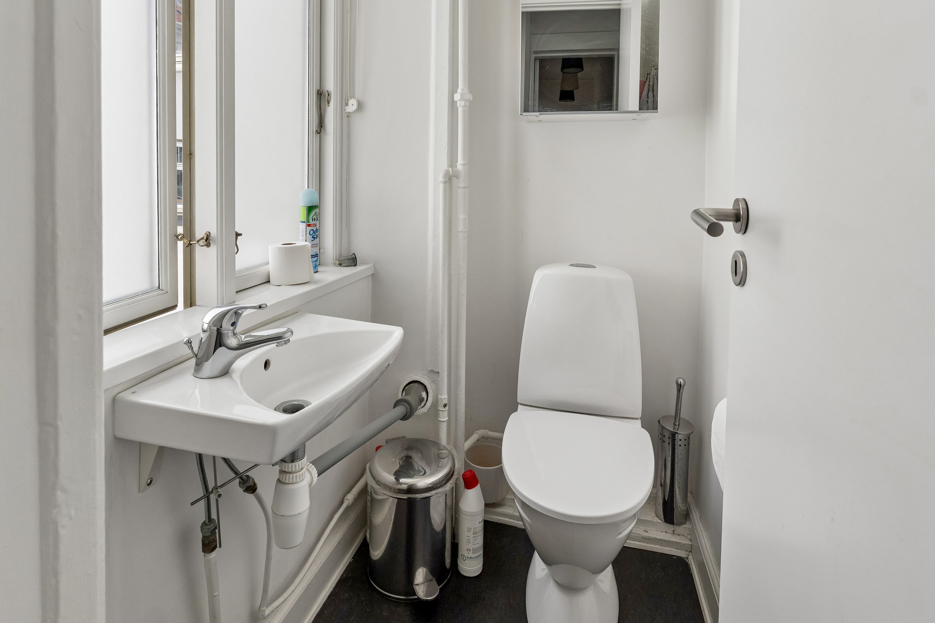 En lille, hvid toiletrum med toilet, håndvask og spildevandsrør samt et toiletpapirhylde og vinduesruder med udsigt til gaden