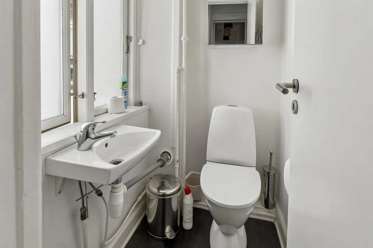 En lille, hvid toiletrum med toilet, håndvask og spildevandsrør samt et toiletpapirhylde og vinduesruder med udsigt til gaden