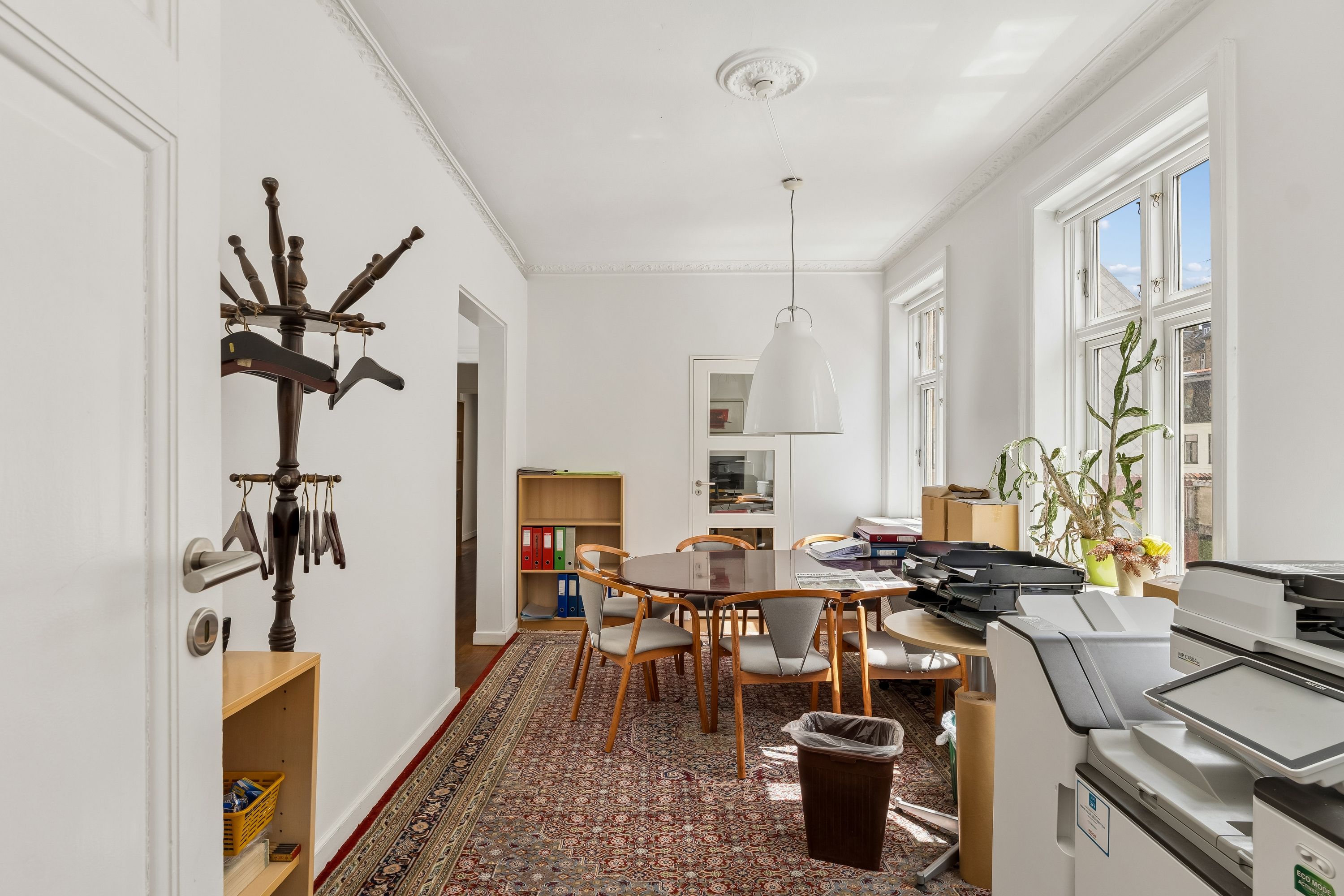 Kontor med skrivebord, stole, plante og papirkurv, skanere og kopierere, møbler og en dekoreret væg.