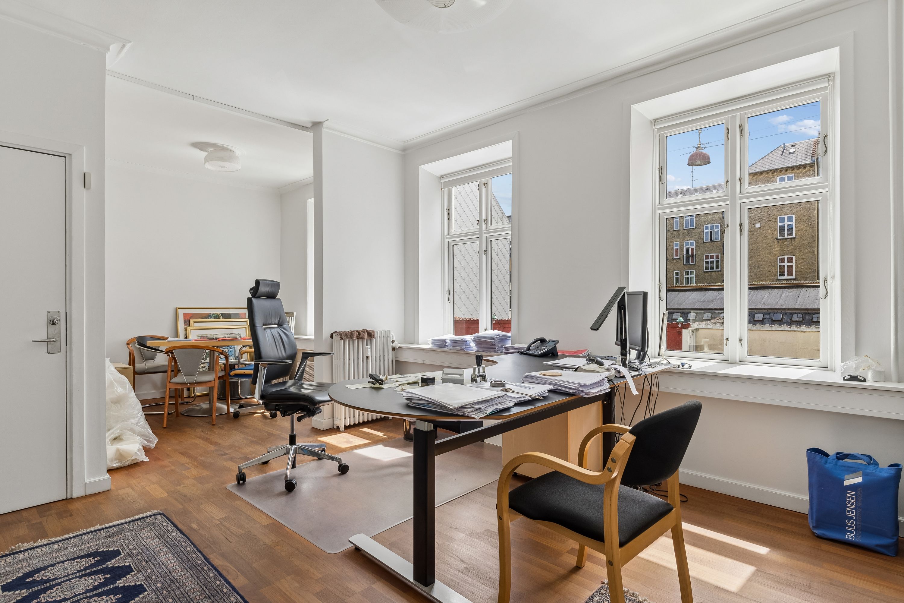 Kontor med bord, stol, computer, to vinduer med udsigt og et køkkenbord med opbevaring i et velmøbleret værelse med lysegule vægge og trægulv