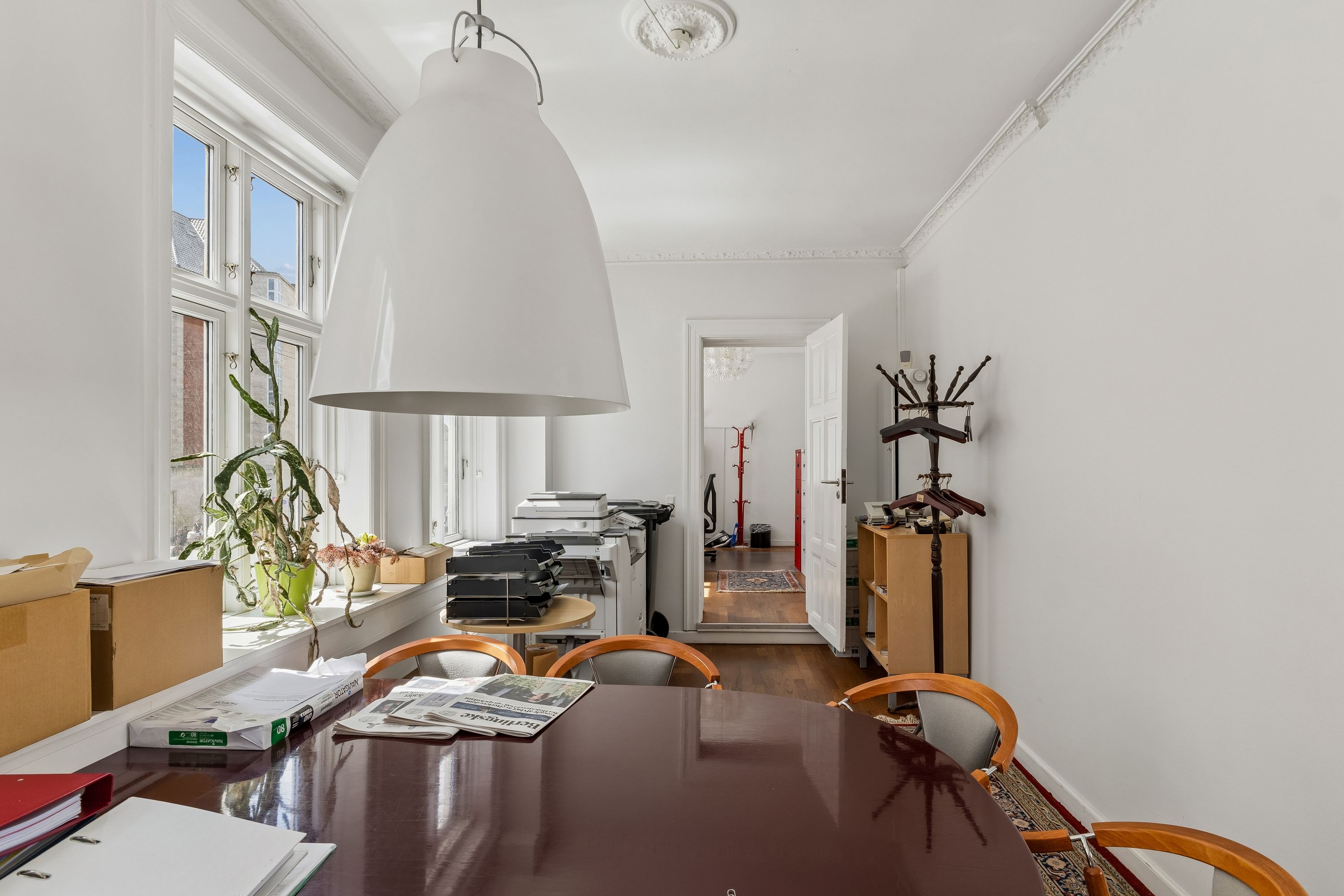 Kontorrum med lyse gulvbelægning og højloft, fuld af møbler og plante. En stor borde med aviser, mapper og en lysekron.
